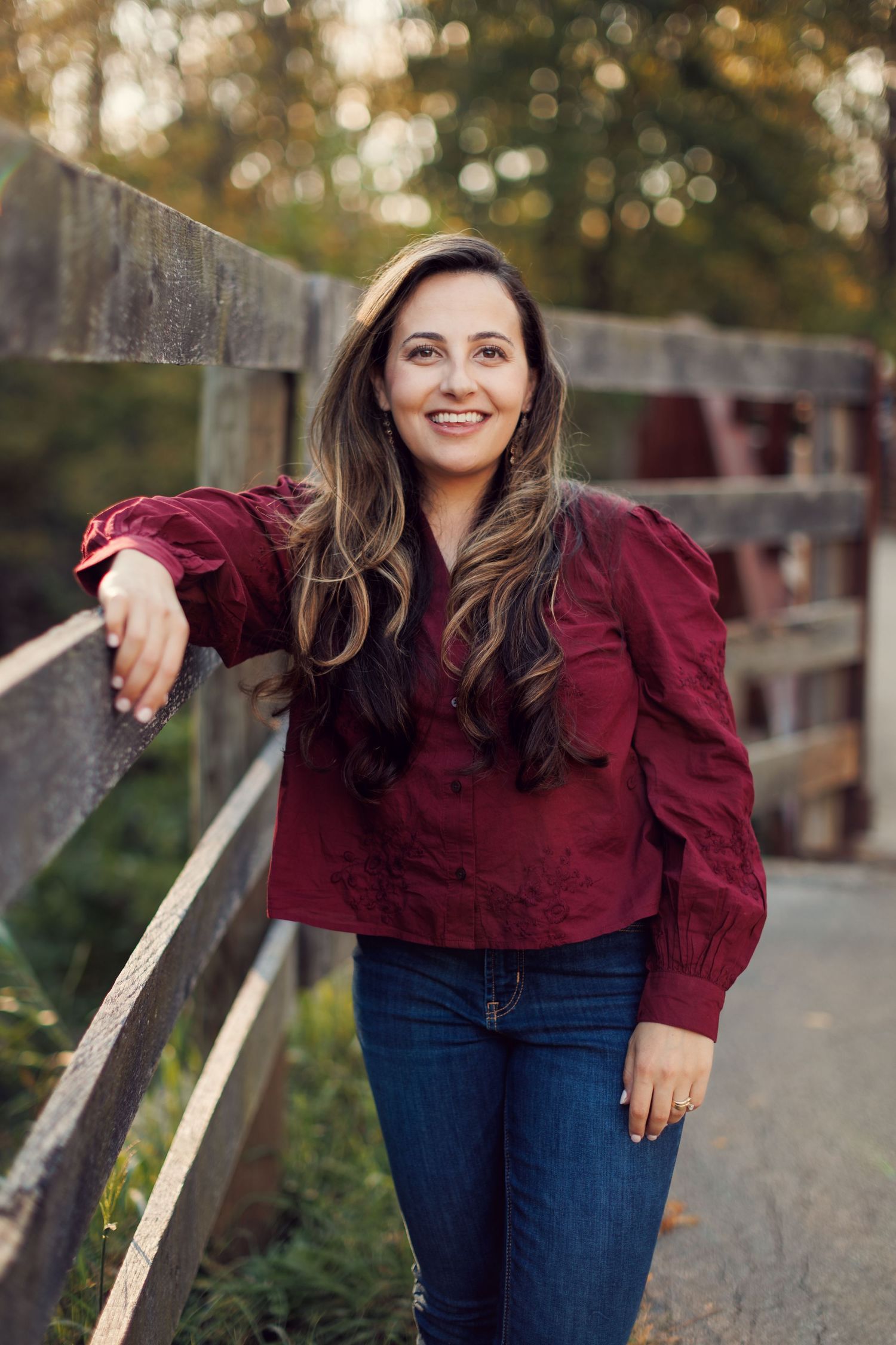 A woman in a burgundy blouse and jeans poses casually against a wooden fence in an outdoor setting.