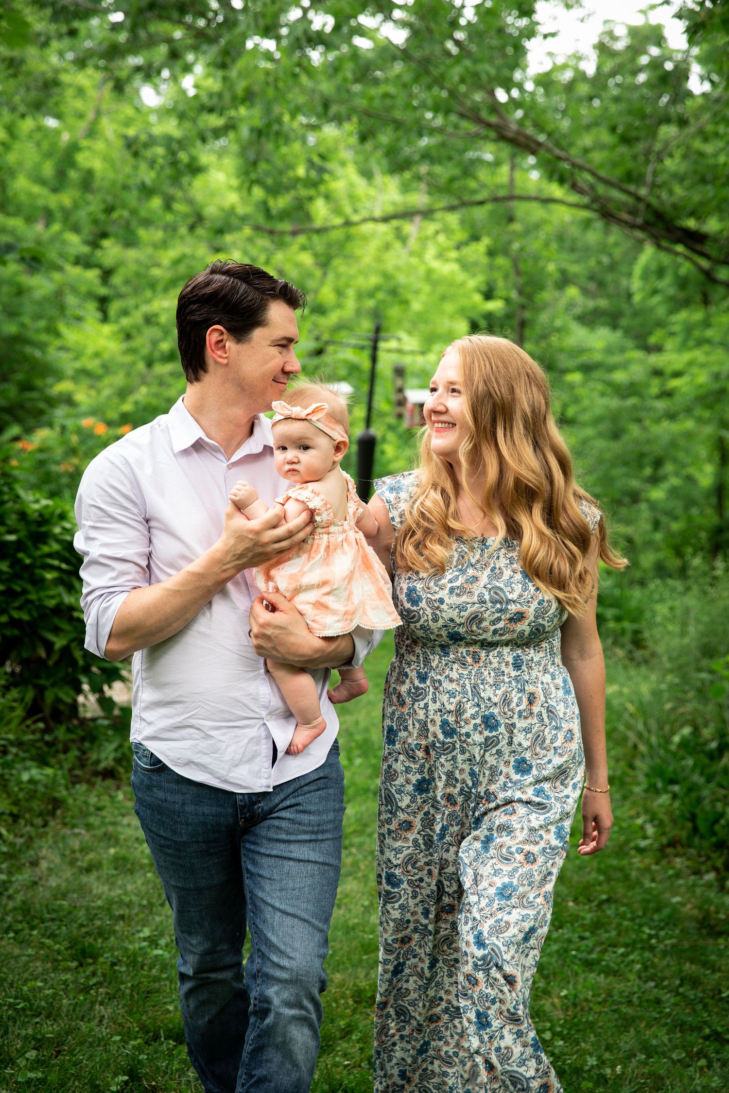 Happy couple in casual dress share a joyful moment with their baby while walking in a lush green forest setting.