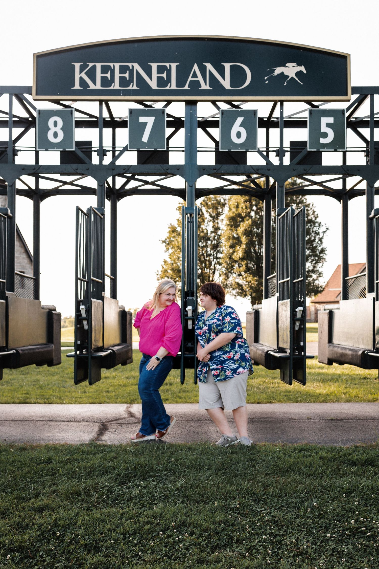 A couple sits under the iconic Keeneland entrance sign against a sunset backdrop for an engagement photo shoot.