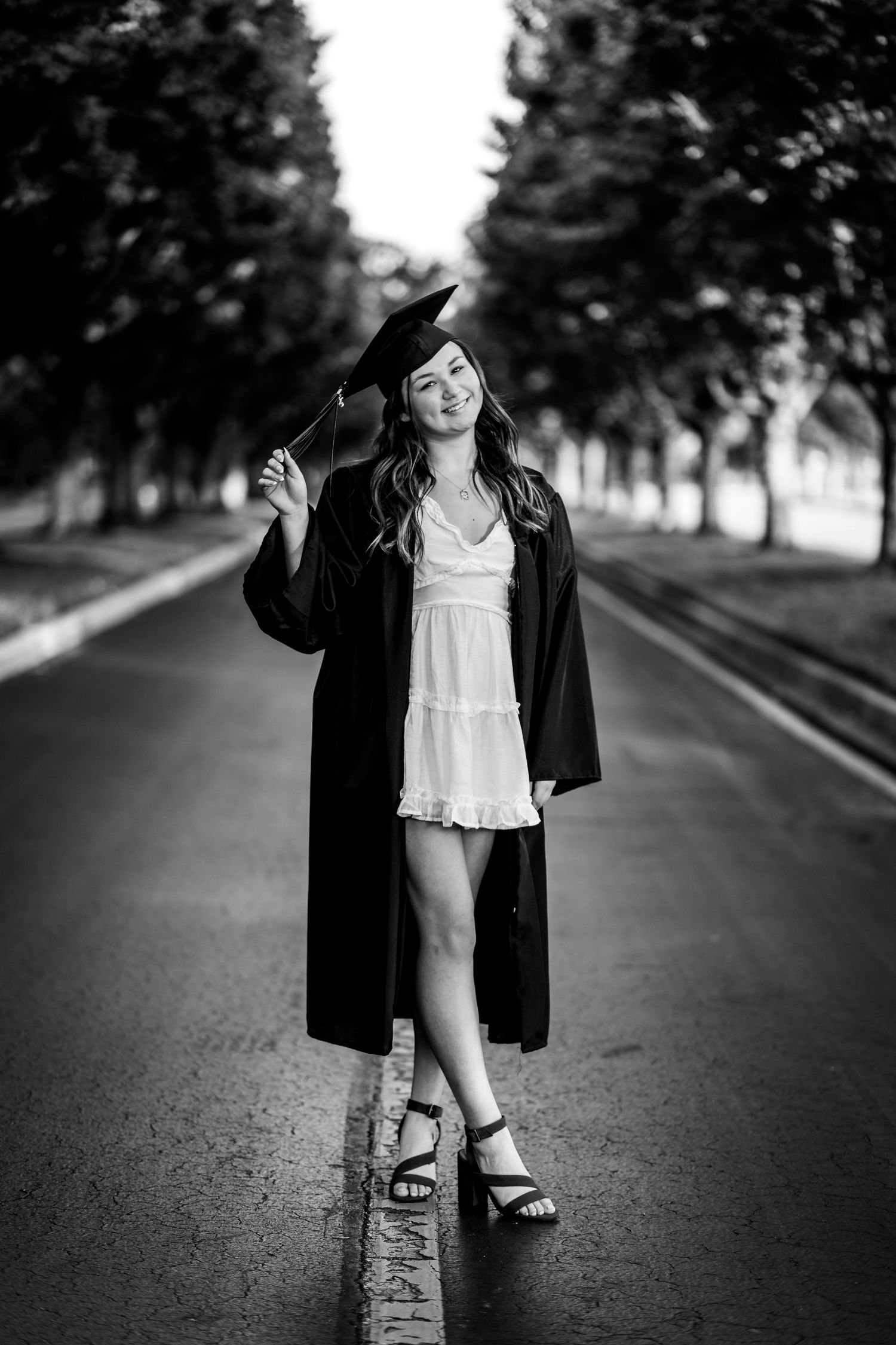 Black and white graduation photo showing graduate in cap and gown walking down tree-lined pathway.