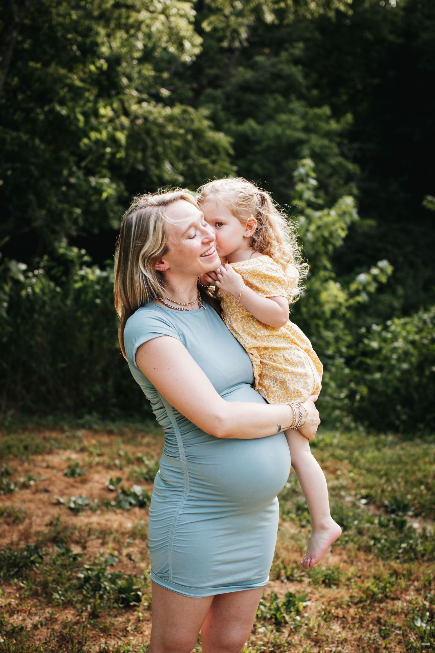A pregnant woman in a blue shirt shares joyful moments with a young child in a natural outdoor setting.
