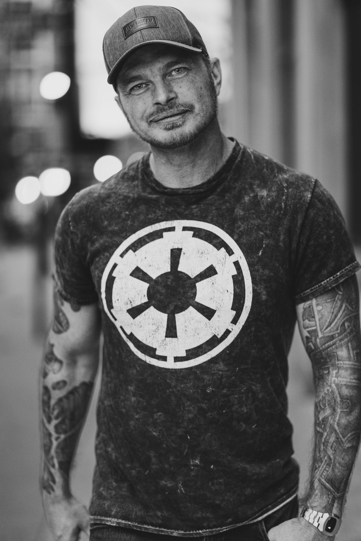 A person wearing a black t-shirt with a white Empire logo from Star Wars shows their tattooed arms in a black and white photo.
