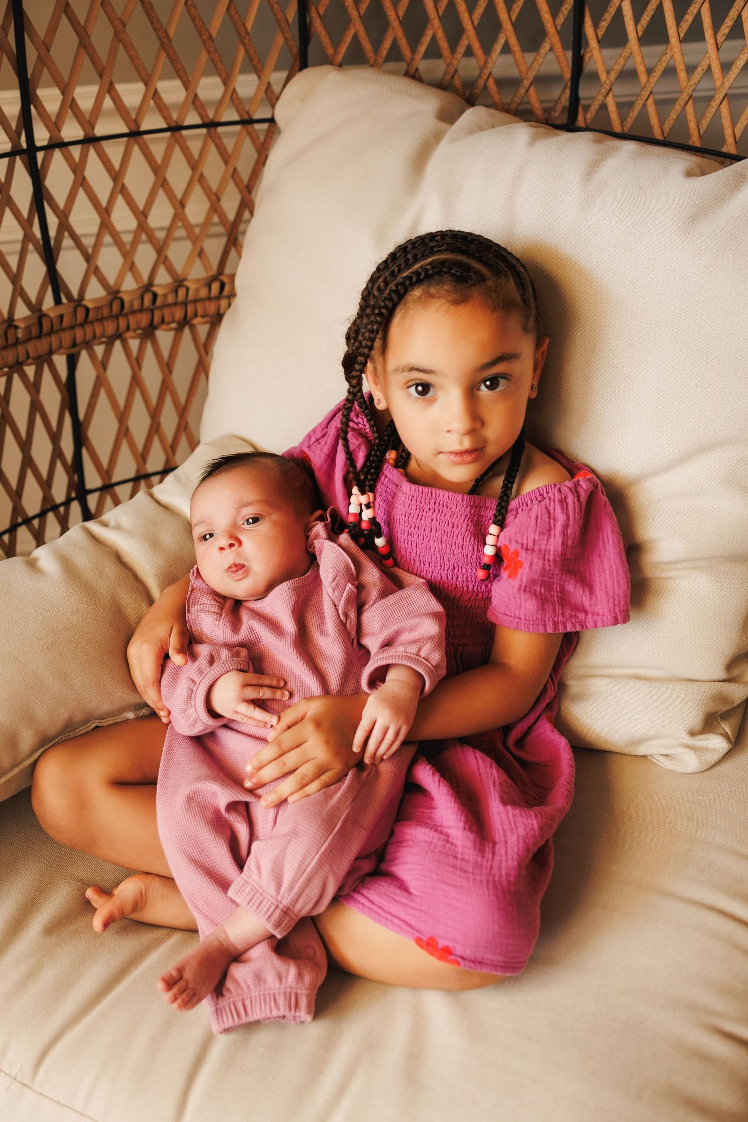 Two children wearing pink outfits resting together on a cream colored couch.