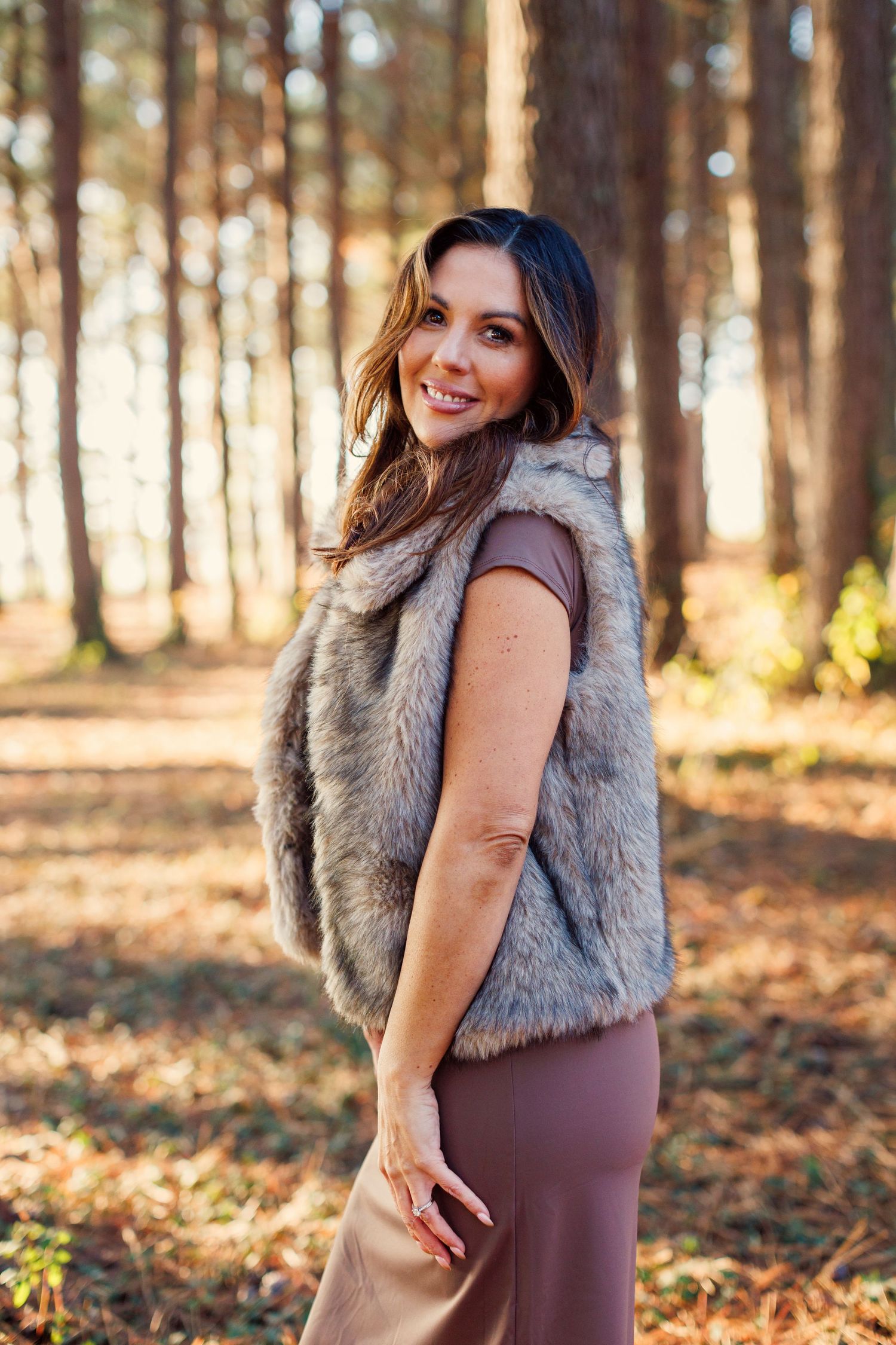 Woman in brown dress and fur vest poses in autumn forest during golden hour sunset.