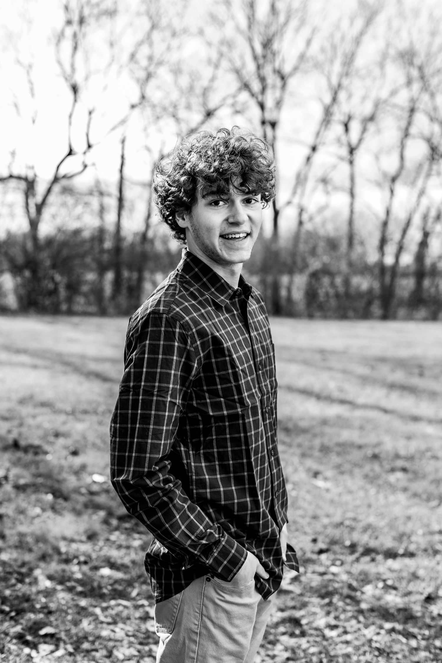 Black and white portrait of someone in plaid shirt smiling against winter trees.