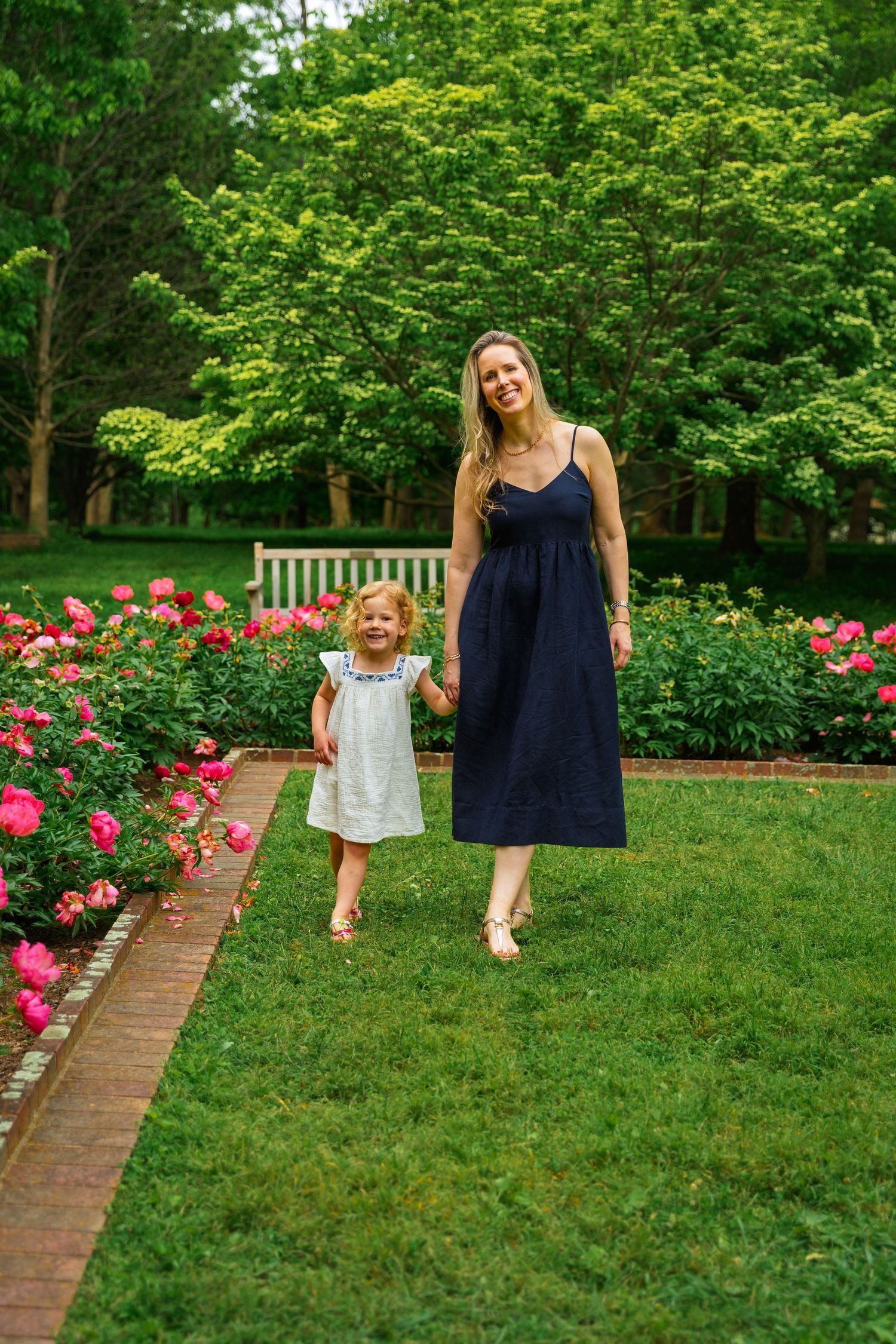 Adult and child walking together on a lush garden path lined with blooming pink roses and green trees.