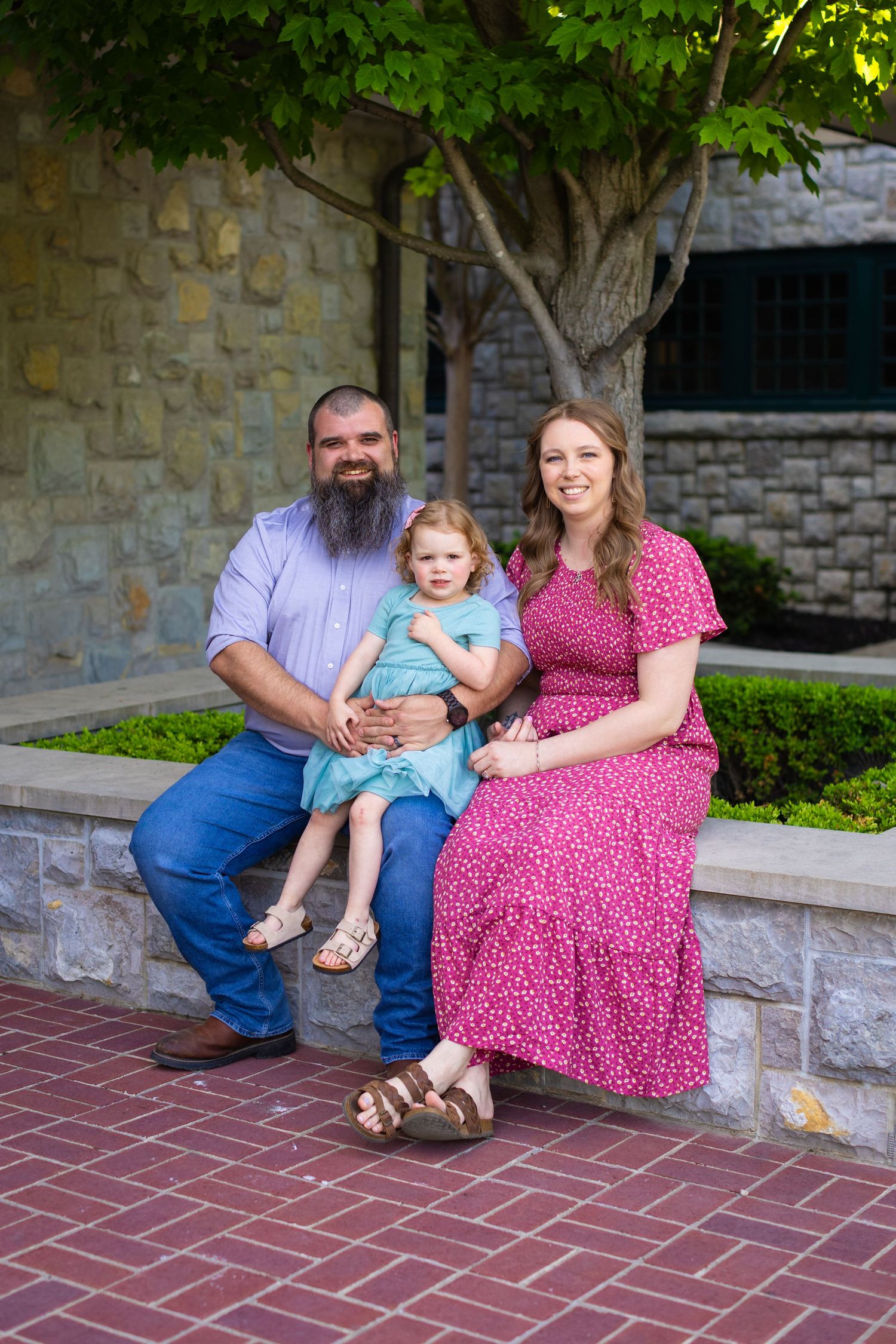 A family sits together on a stone ledge near a brick path and stone wall with green trees above.