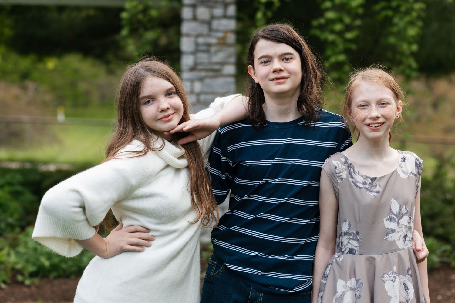 Three siblings pose together outdoors against a stone wall and garden backdrop.