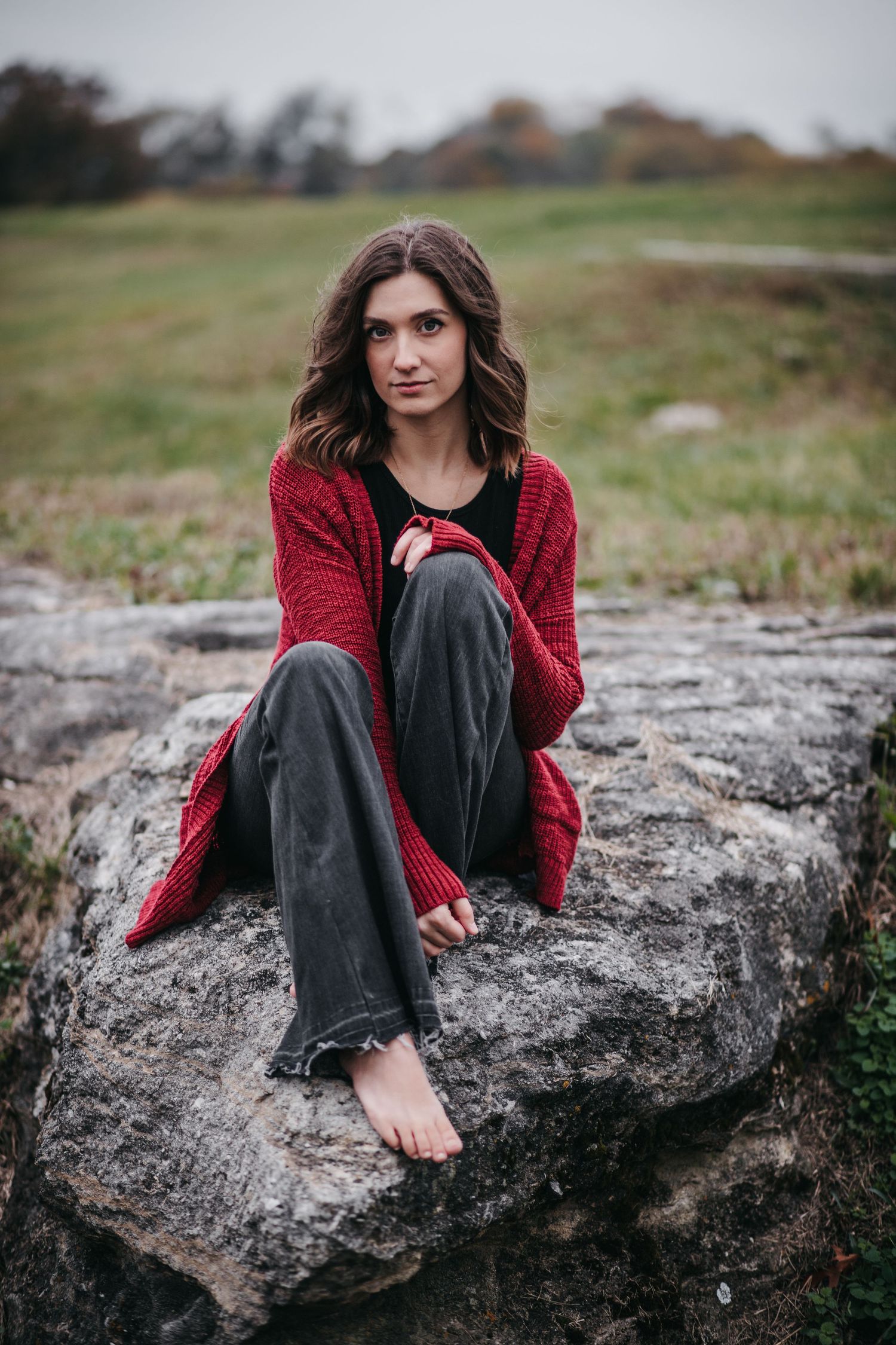 Portrait of person in red cardigan sitting barefoot on rocky outcrop against moody autumn landscape.
