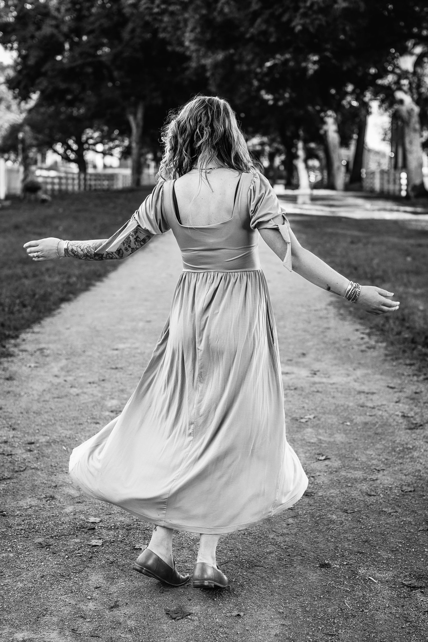 A person in a flowing white dress twirls gracefully on a tree-lined path in black and white photography.