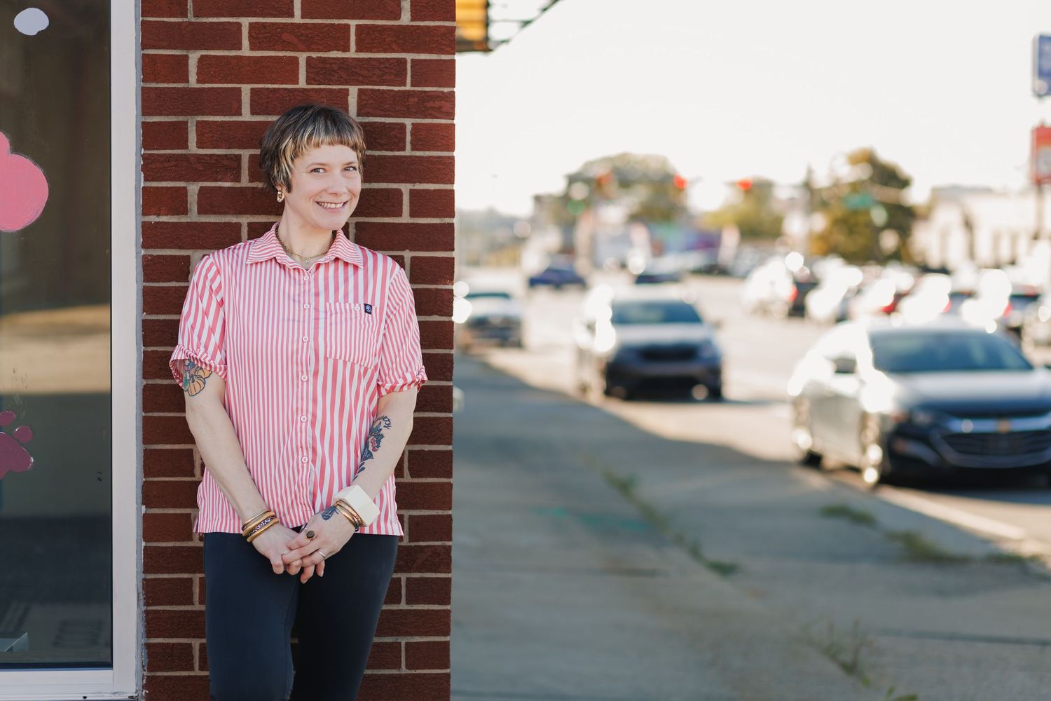A person in a pink striped shirt stands against a brick wall in a parking lot.