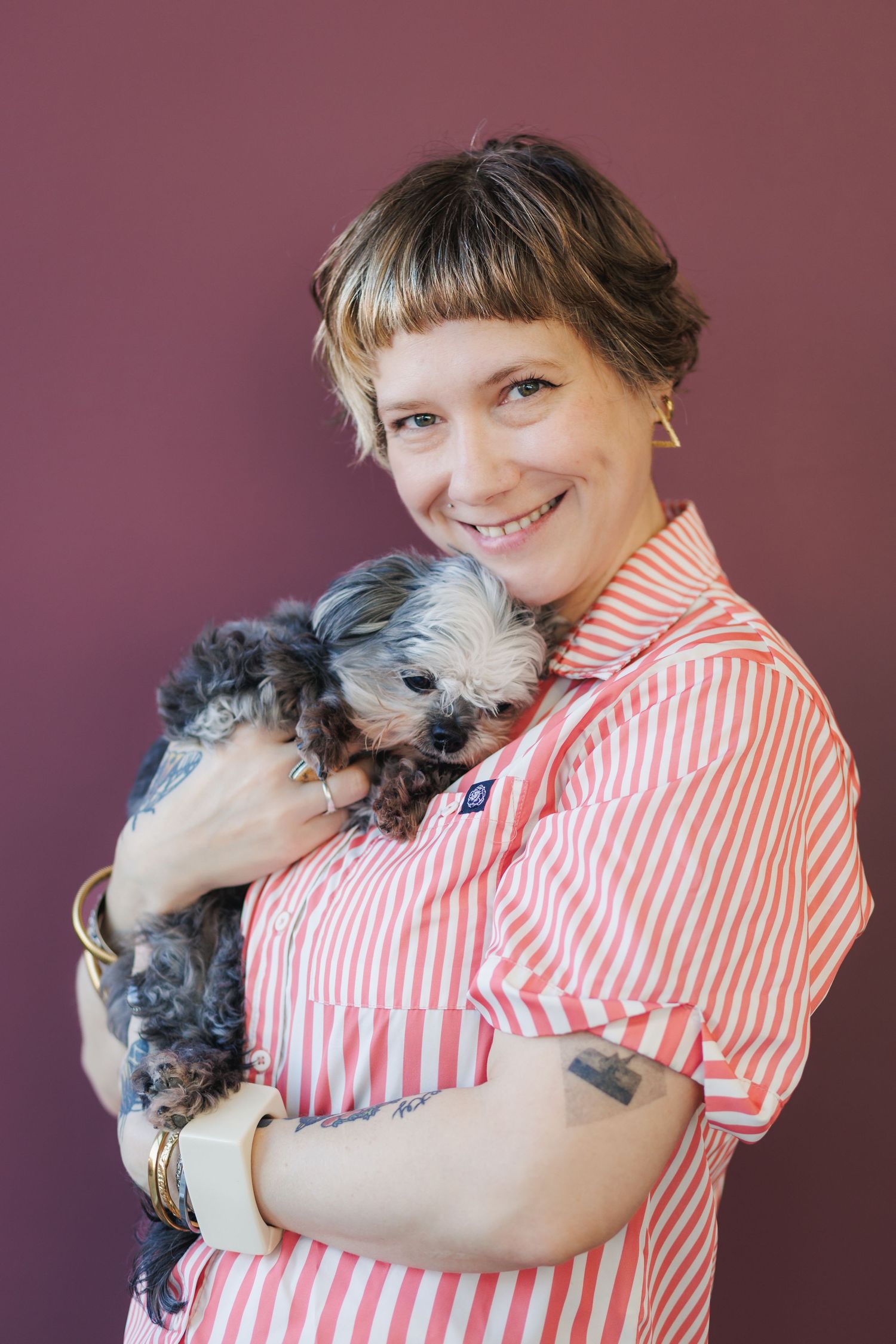 A person in a red and white striped shirt hugs and snuggles with a small gray scruffy dog against a burgundy background.