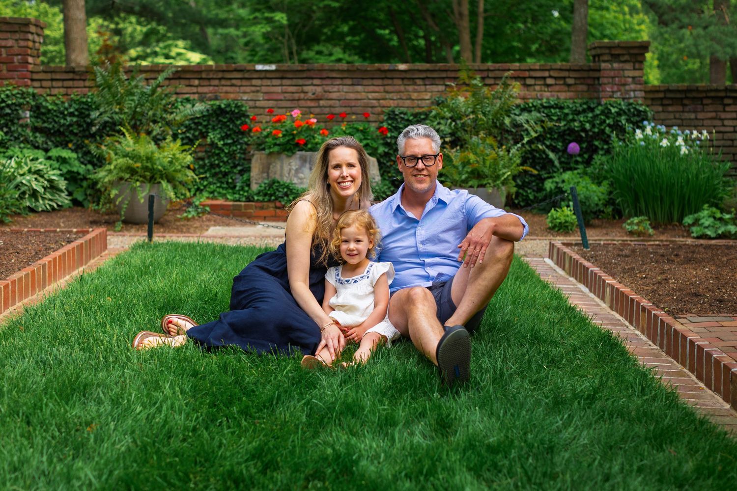 Family enjoys a relaxing moment together on lush green grass in a backyard garden setting with brick landscaping.