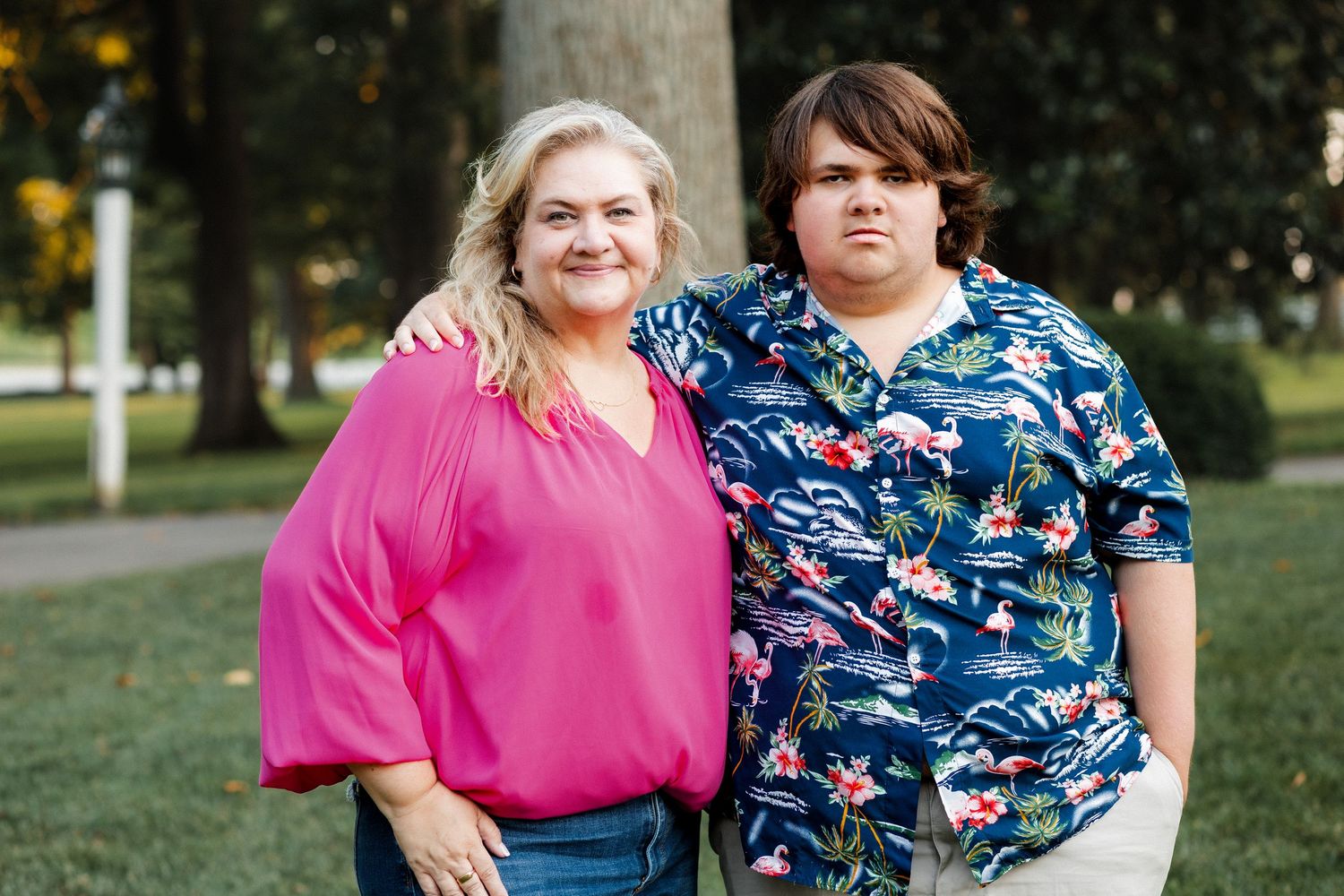 Two people stand together outdoors wearing casual attire with a pink blouse and blue patterned shirt against trees.