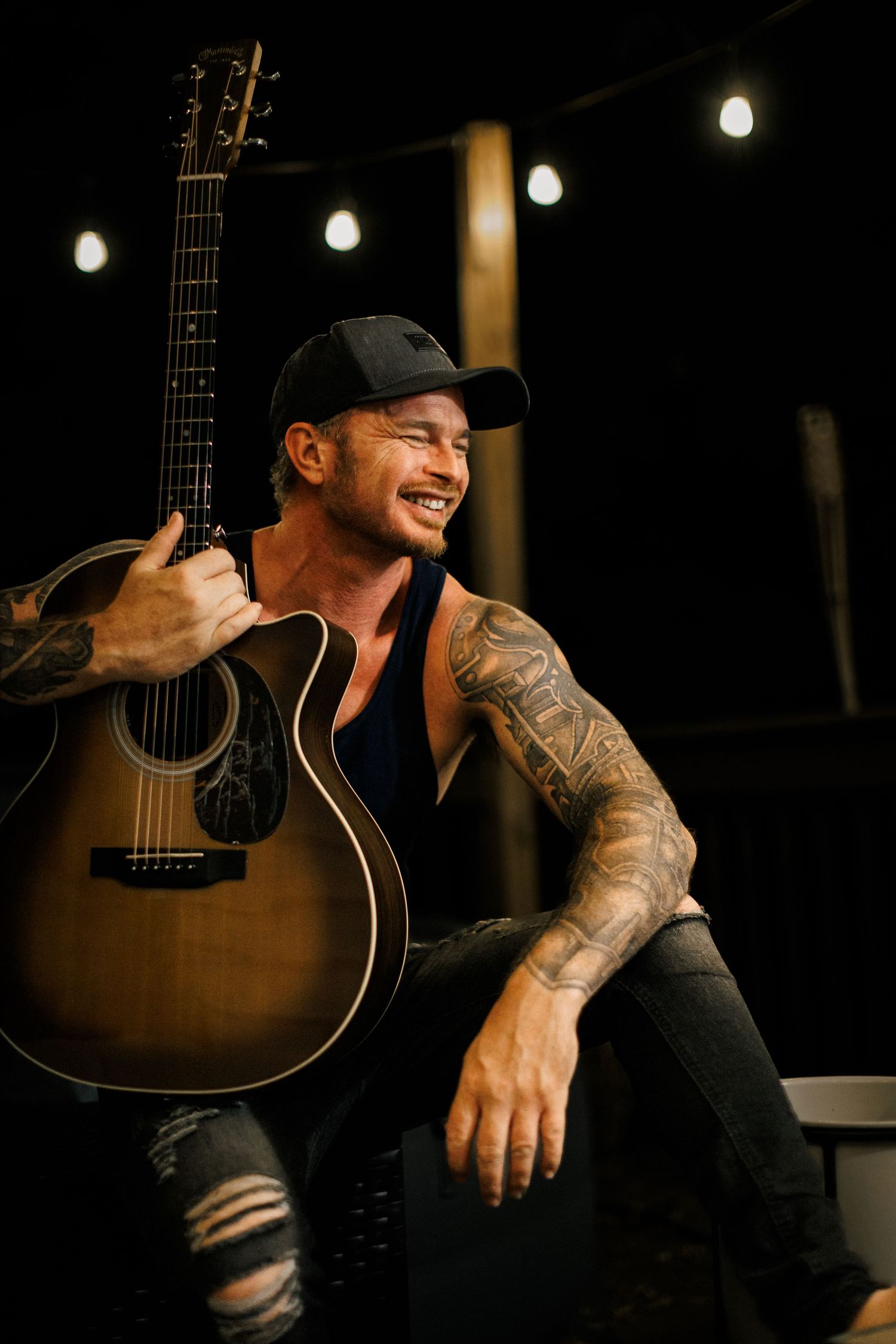 A performer in black clothing and baseball cap sits with an acoustic guitar in a dimly lit setting with string lights.