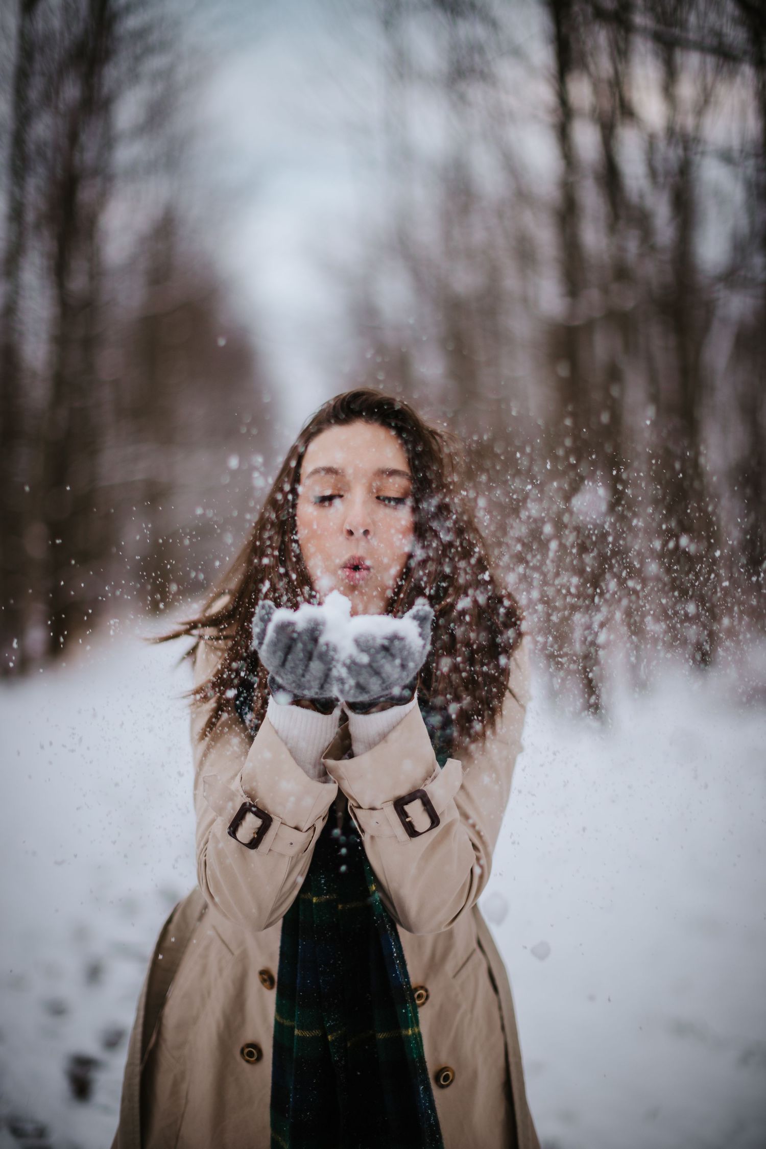 Person in beige coat blowing snow from gloved hands in magical winter forest setting.