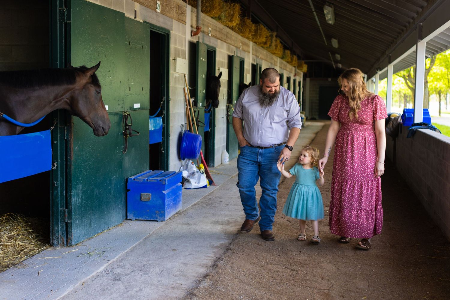 People walk past horse stables with a brown horse peeking out from a stall in the background.