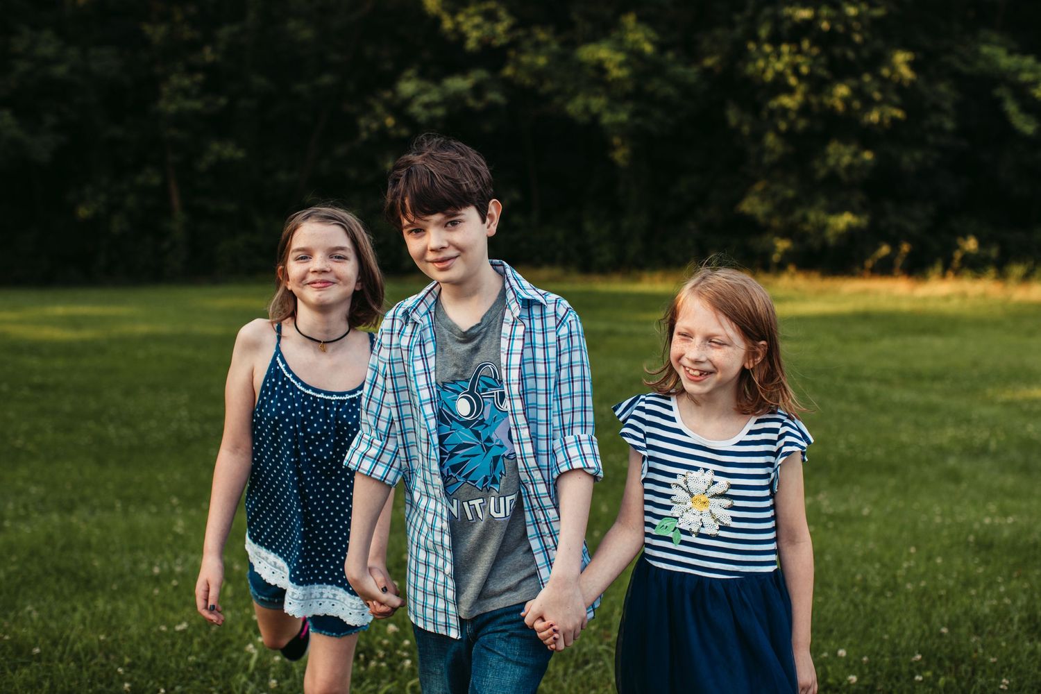 Three children in casual clothing walk together and smile in a sunny outdoor setting.