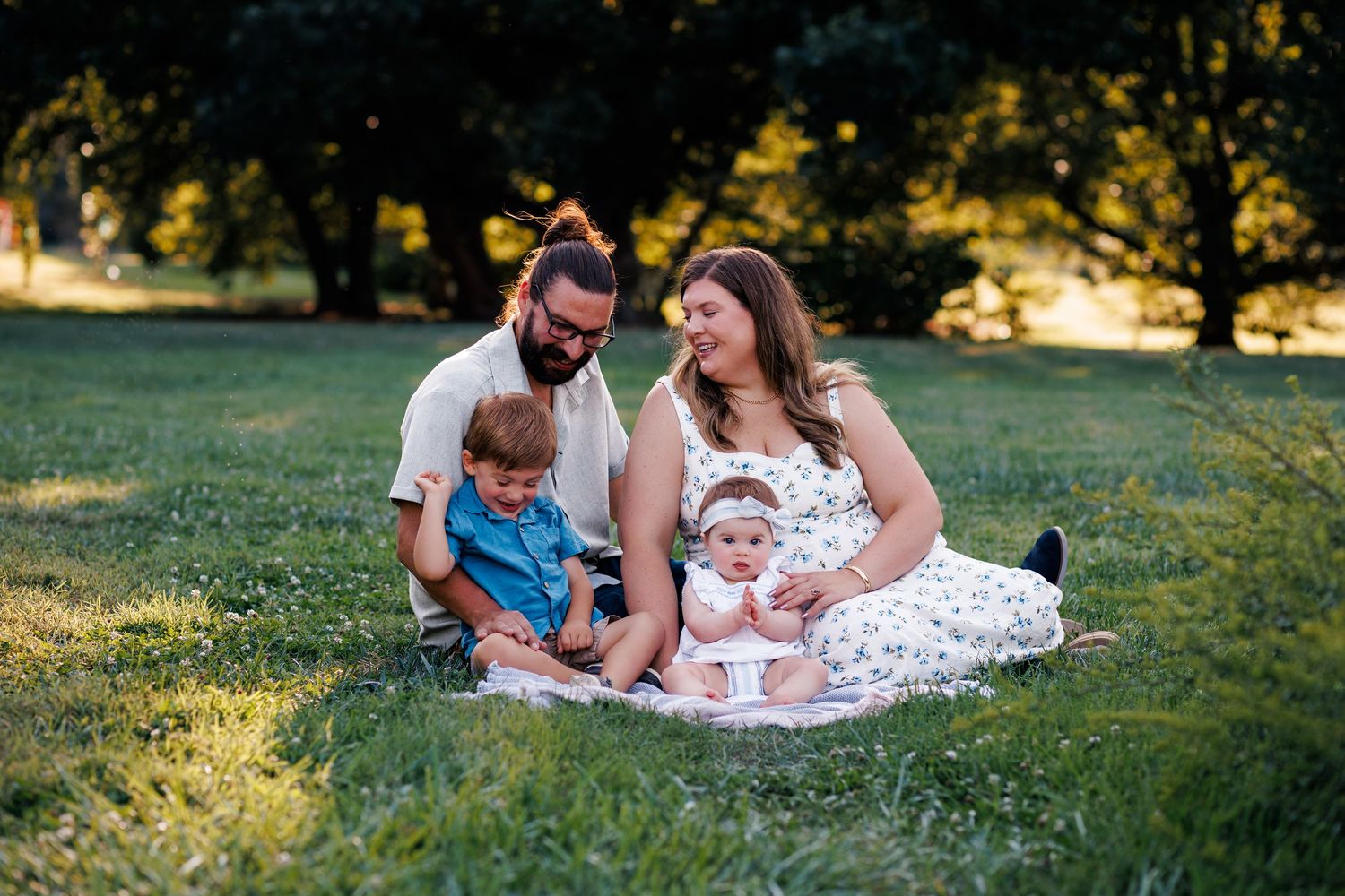Family sits together on picnic blanket in grassy park during golden hour sunset.