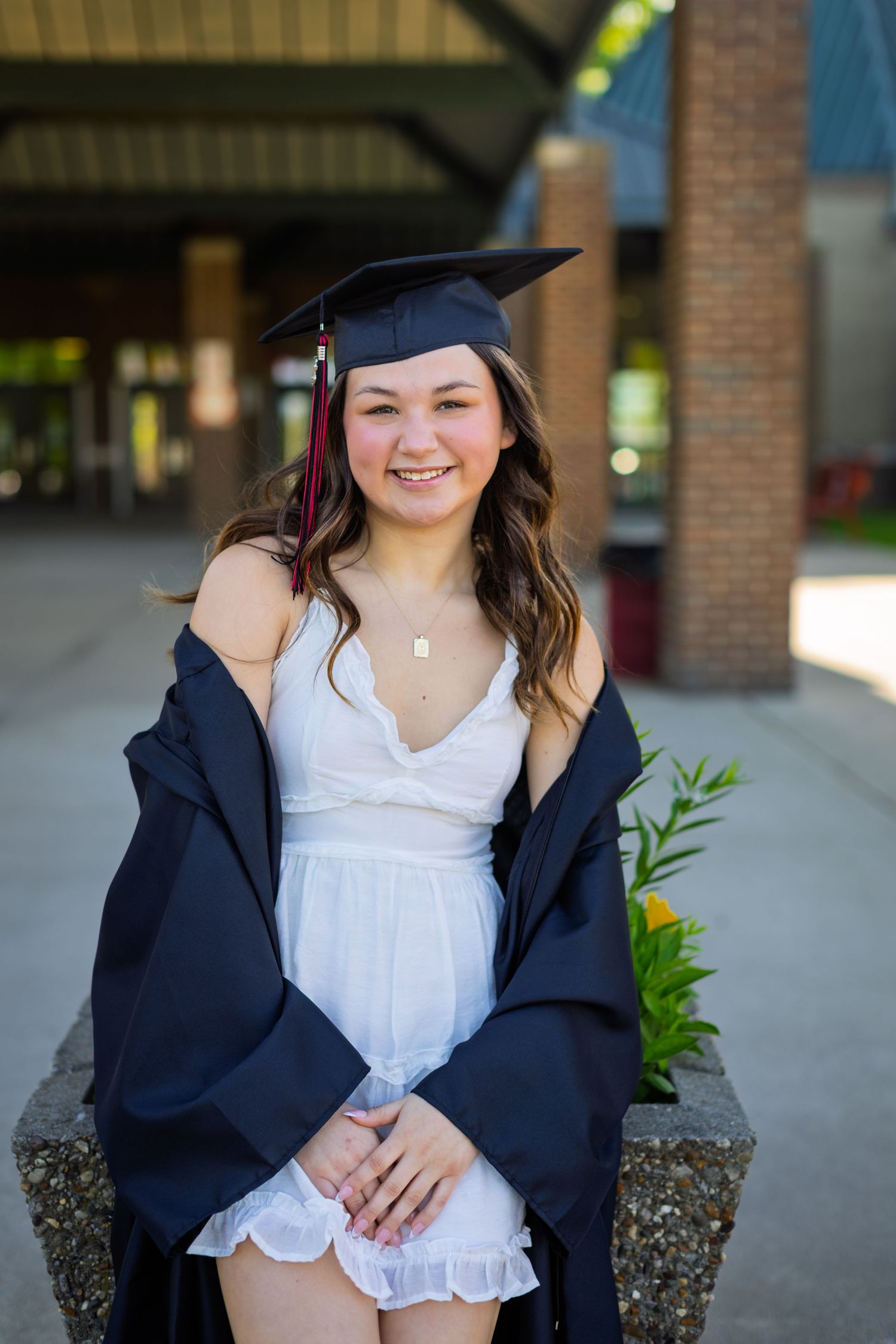 Graduate in cap and gown wearing white dress sits on concrete ledge during outdoor graduation photo session.
