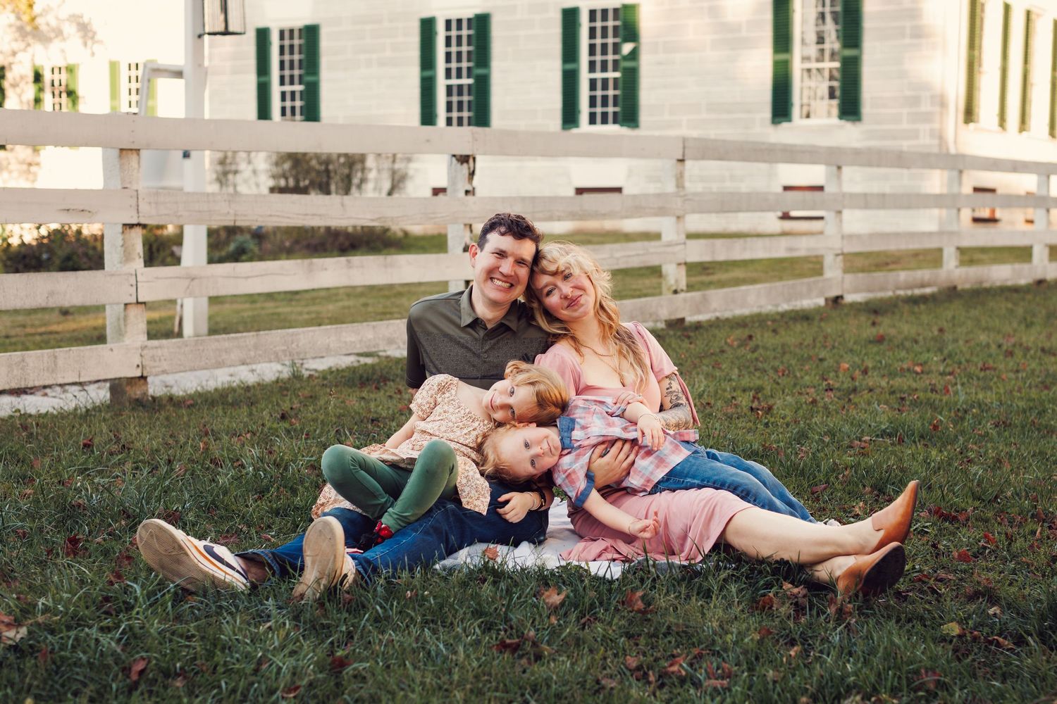 Couple relaxing on grass by white fence during sunset engagement photo session.