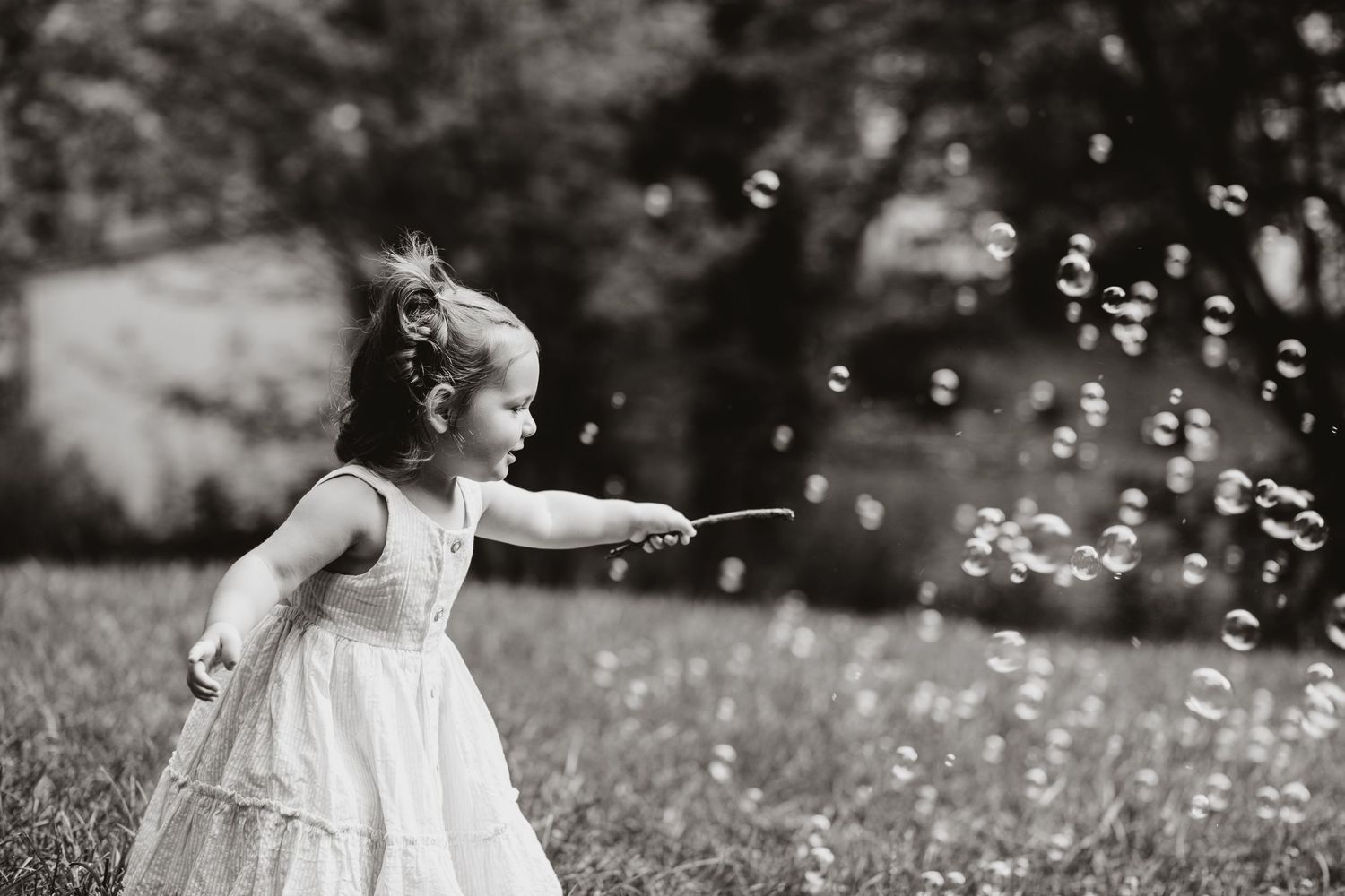 Black and white image of a child in a white dress chasing bubbles in a meadow.