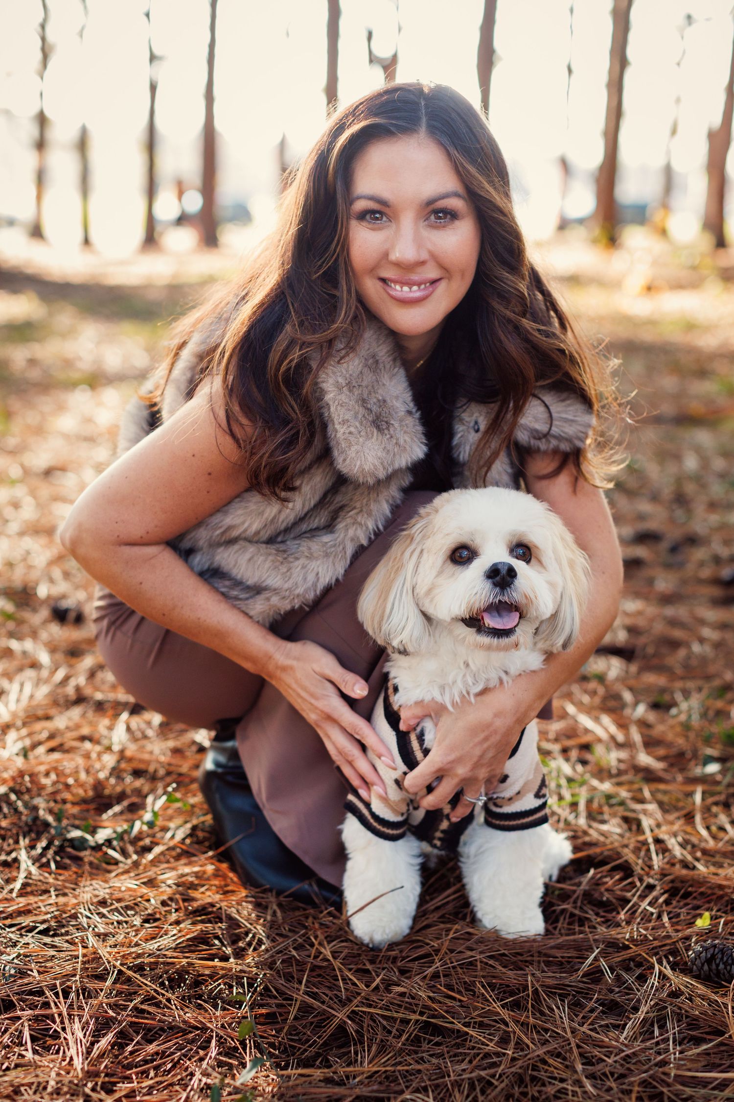 A small white Shih Tzu puppy sits in autumn leaves with someone wearing a brown sweater and fur vest.
