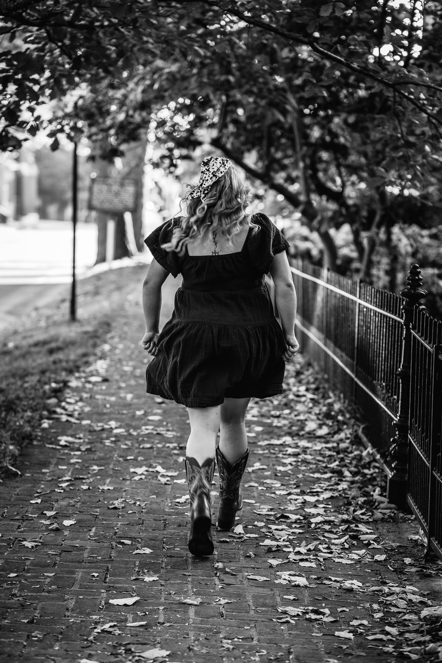 Black and white photo of someone walking on a leaf-covered brick path wearing boots and a dress.