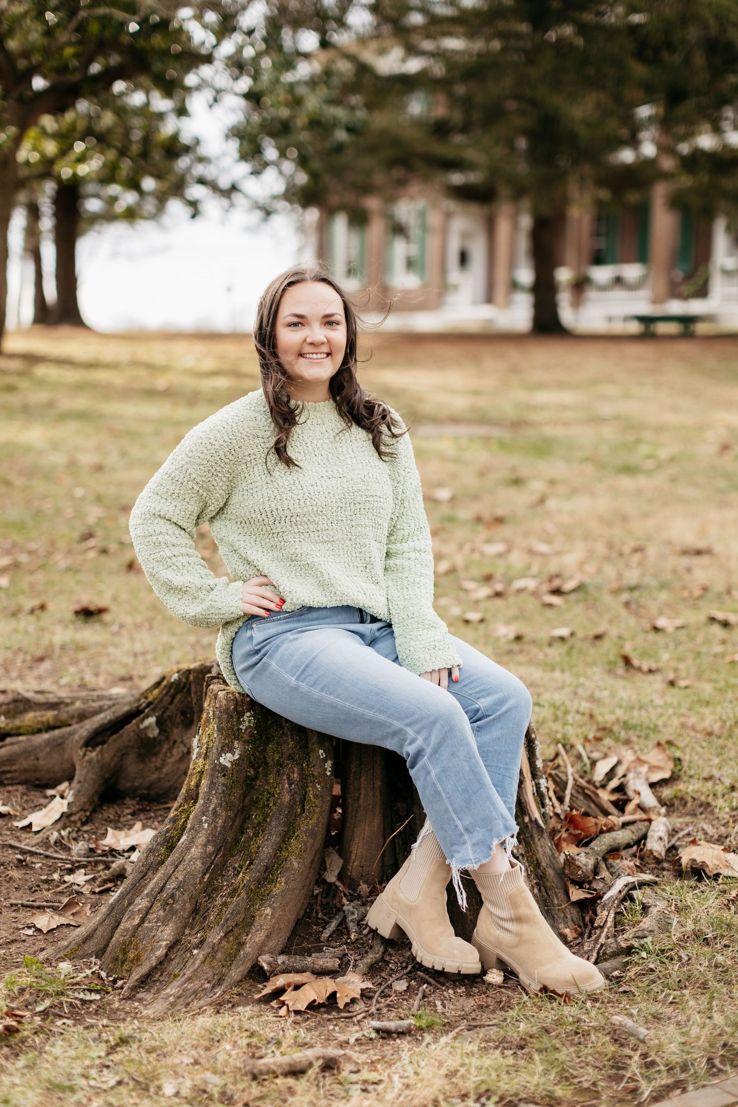 Casual portrait of someone in cream sweater and jeans sitting on tree stump in autumn setting.