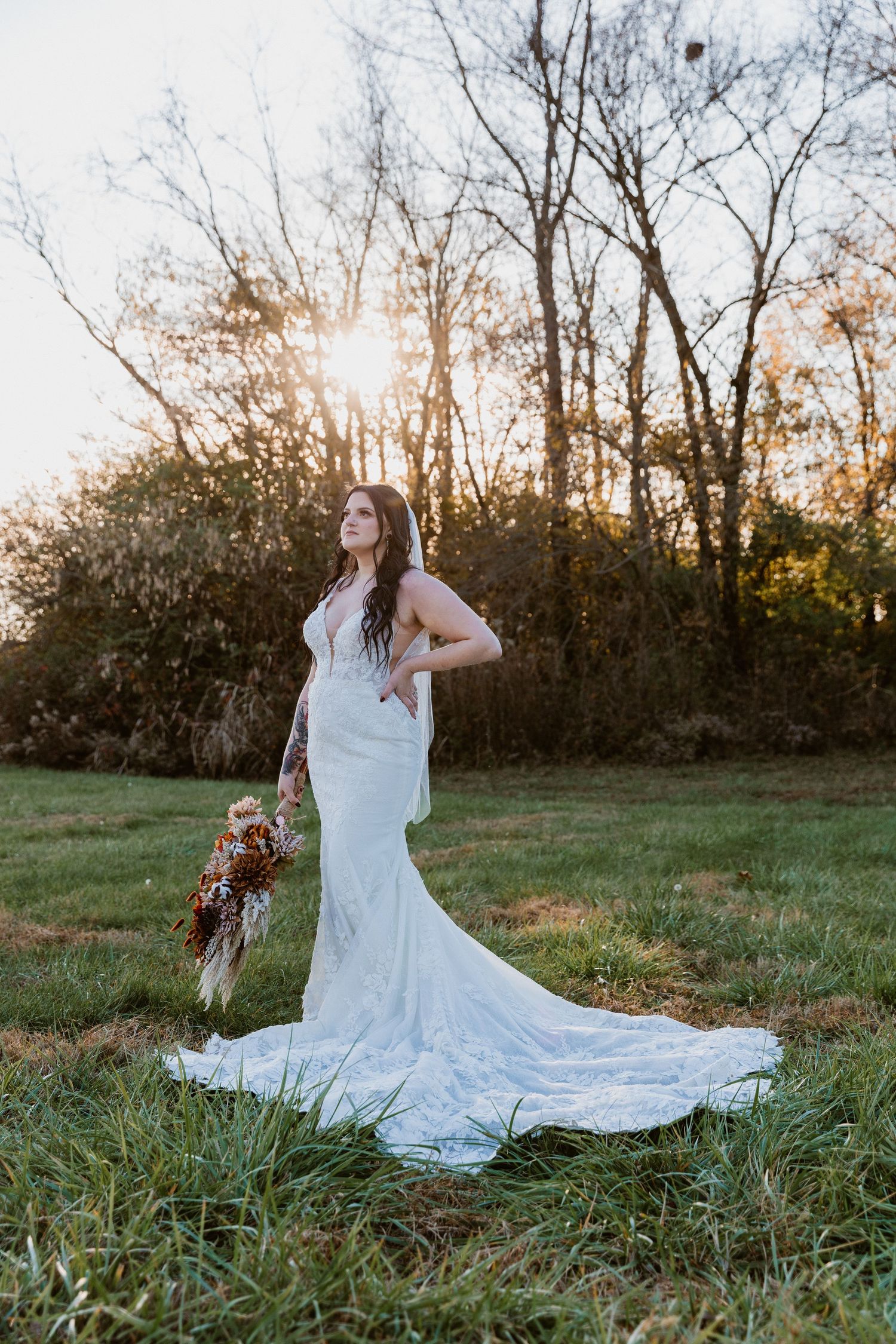 Bride in elegant lace wedding dress poses in field at sunset holding autumn bouquet.