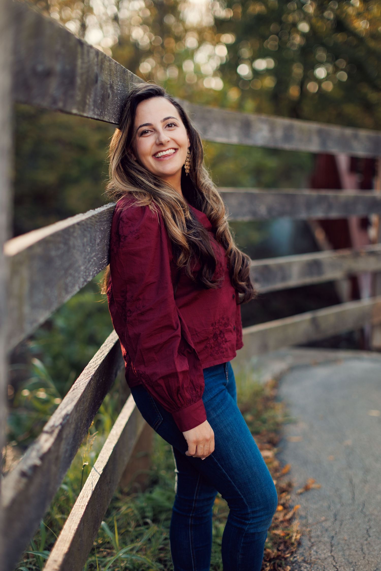 A person in a burgundy sweater and blue jeans smiles while leaning against a wooden fence in natural outdoor lighting.