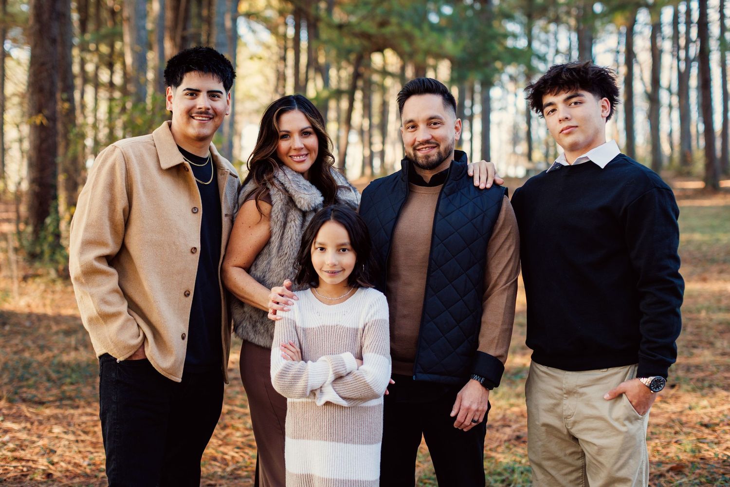 A family poses together outdoors in casual fall attire among tall pine trees and autumn leaves on the ground.