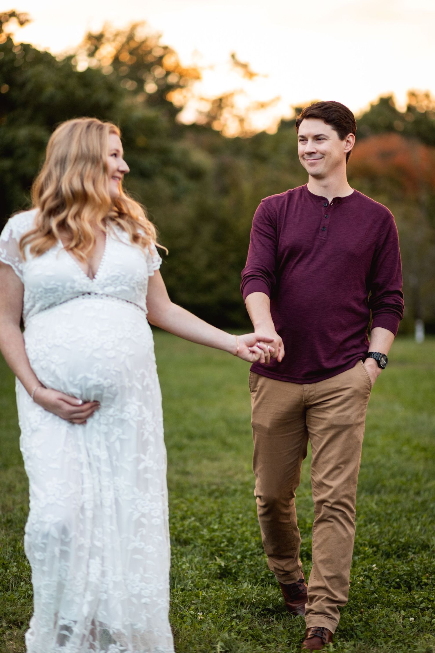 Expectant couple holds hands while walking on grass during golden hour photography session.