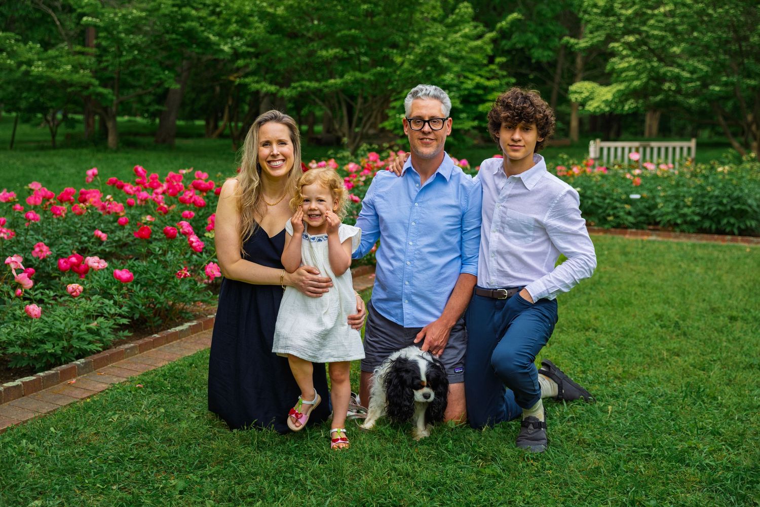 A family and their dog pose together in a garden with blooming pink roses and lush greenery in the background.