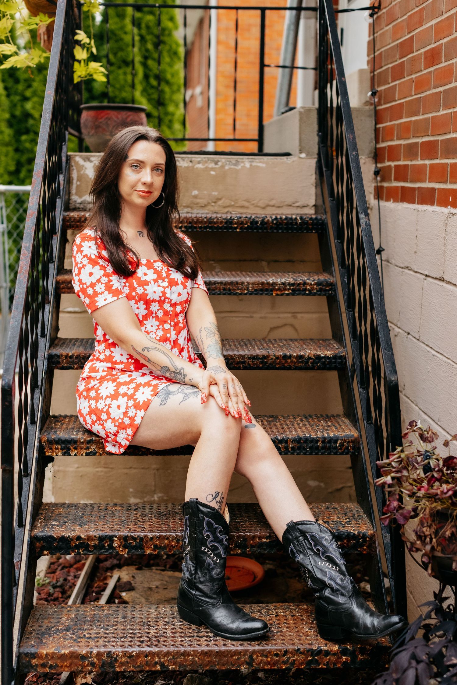 A figure in a red floral dress and black cowboy boots sits on outdoor metal stairs near a brick wall.