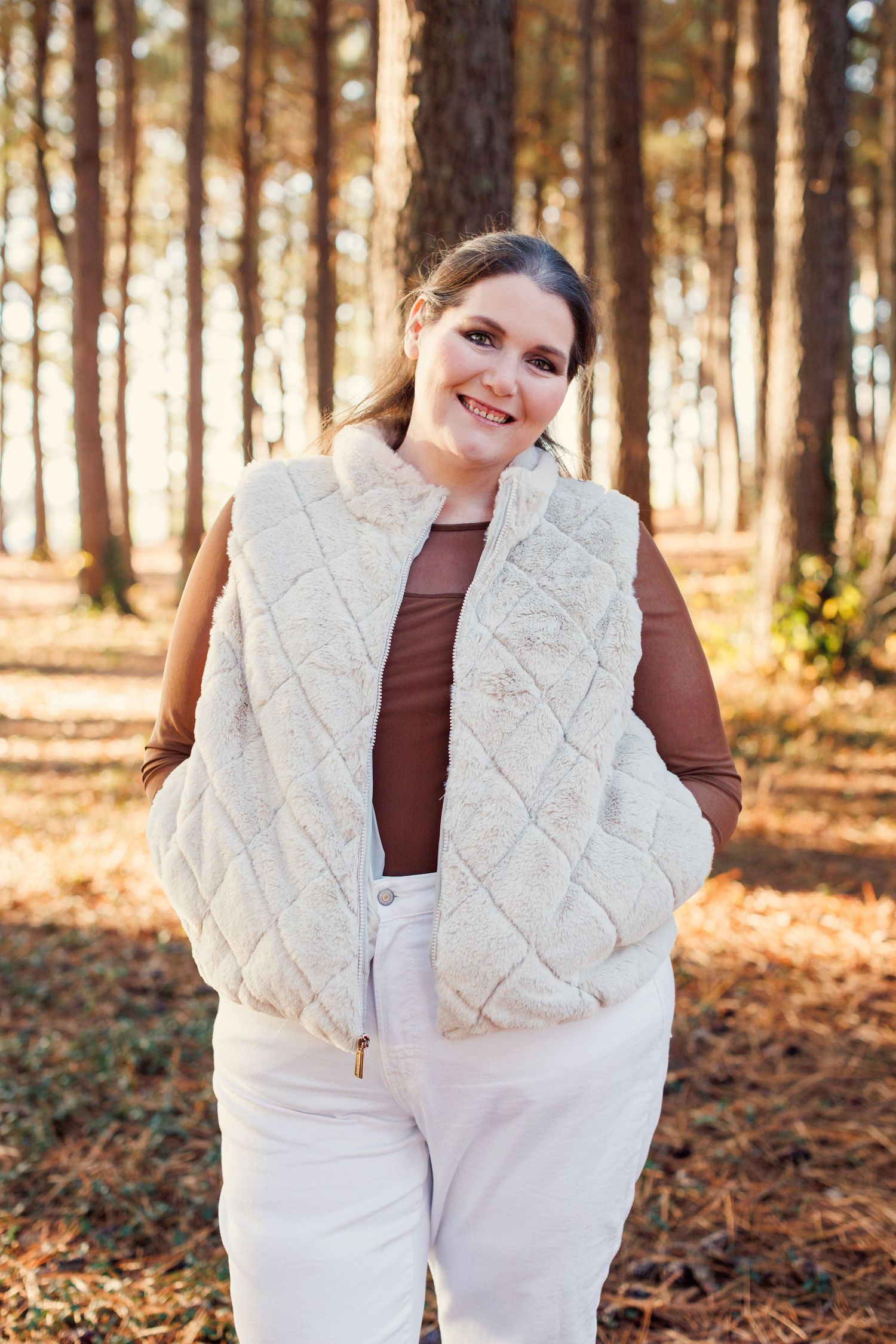 A person wearing a white quilted vest and brown shirt poses in a sunlit autumn forest setting.