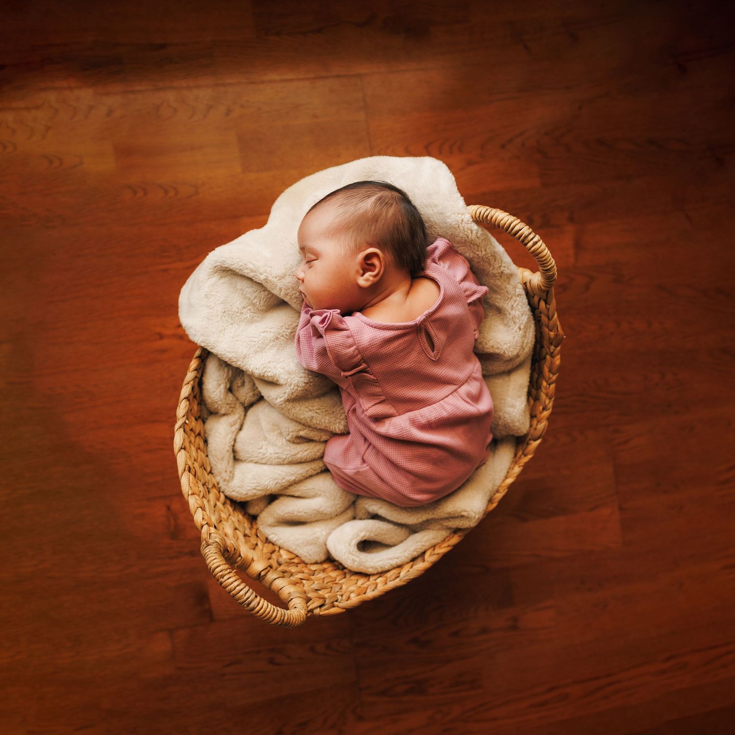 A sleeping baby wearing pink lies curled up in a cozy woven basket with soft white blankets on a wooden floor.