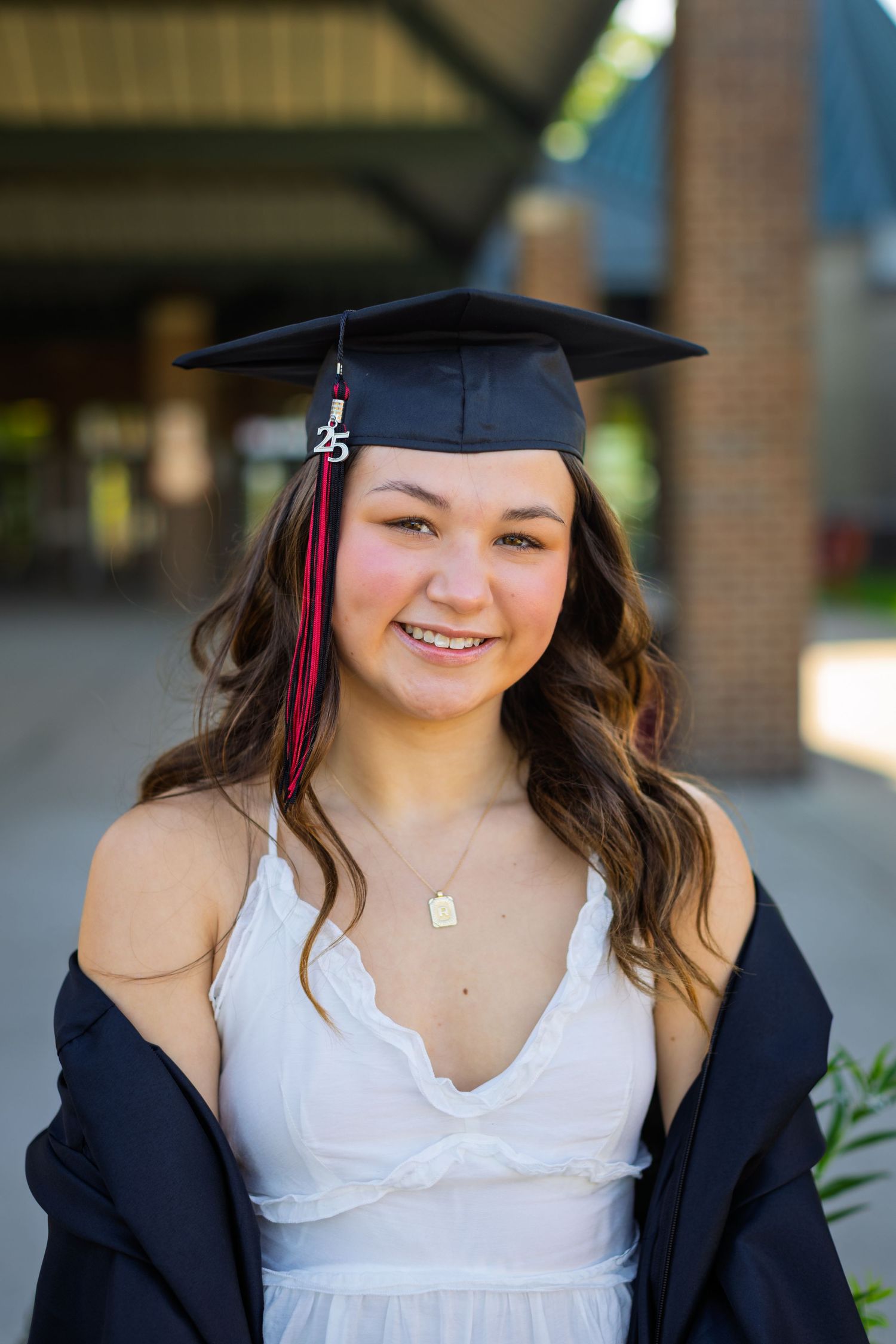 Graduate wearing black cap and gown with white sundress and necklace poses for celebratory photos outdoors.