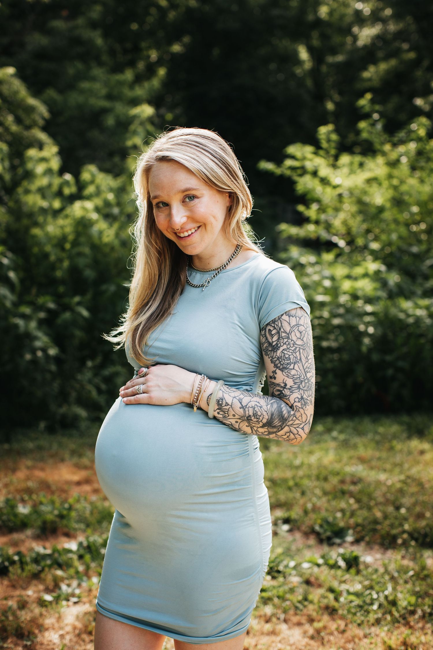 A pregnant woman in a light blue dress poses outdoors with tattoo sleeves against a green background.