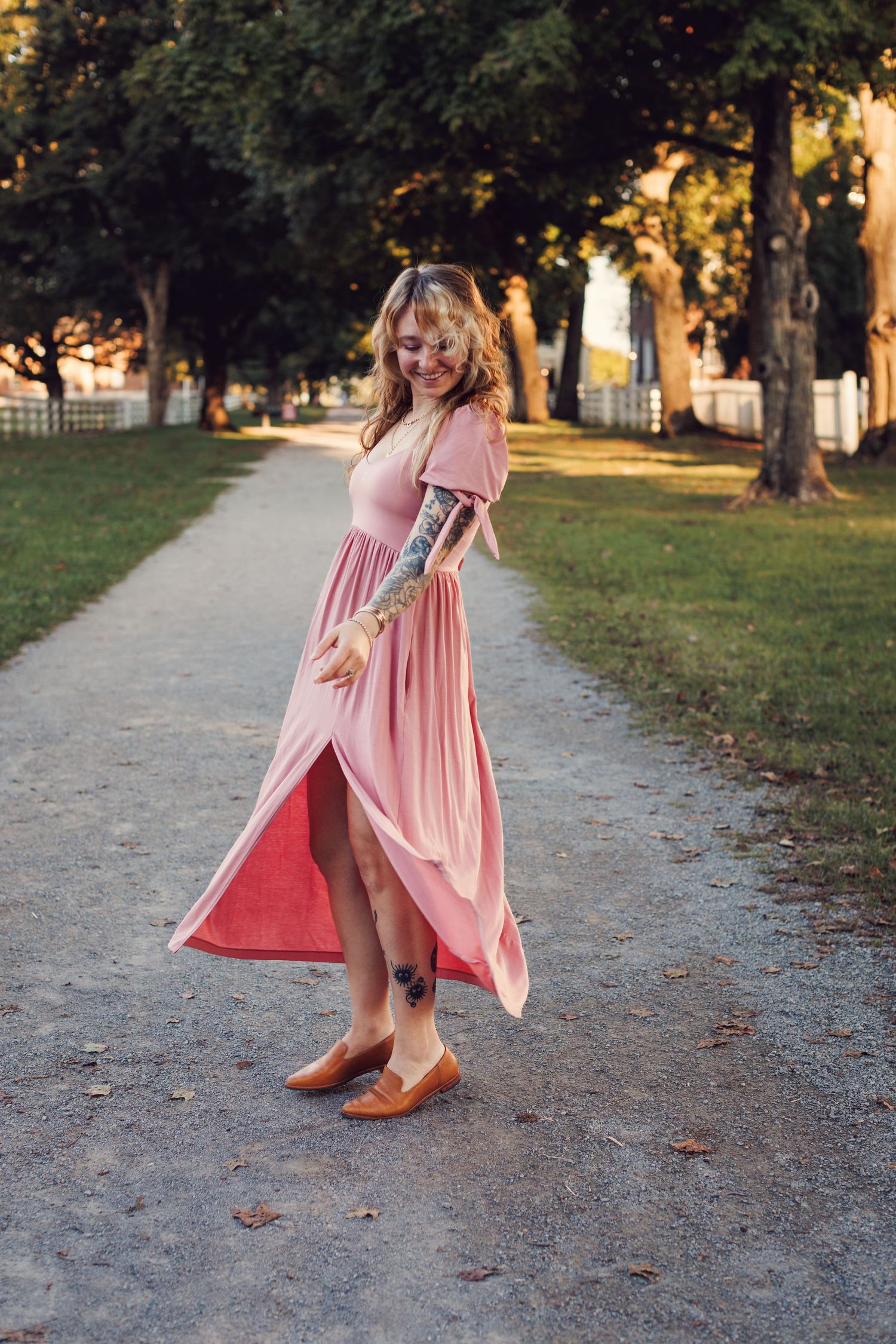 A flowing pink dress twirls gracefully on a tree-lined path during golden hour.