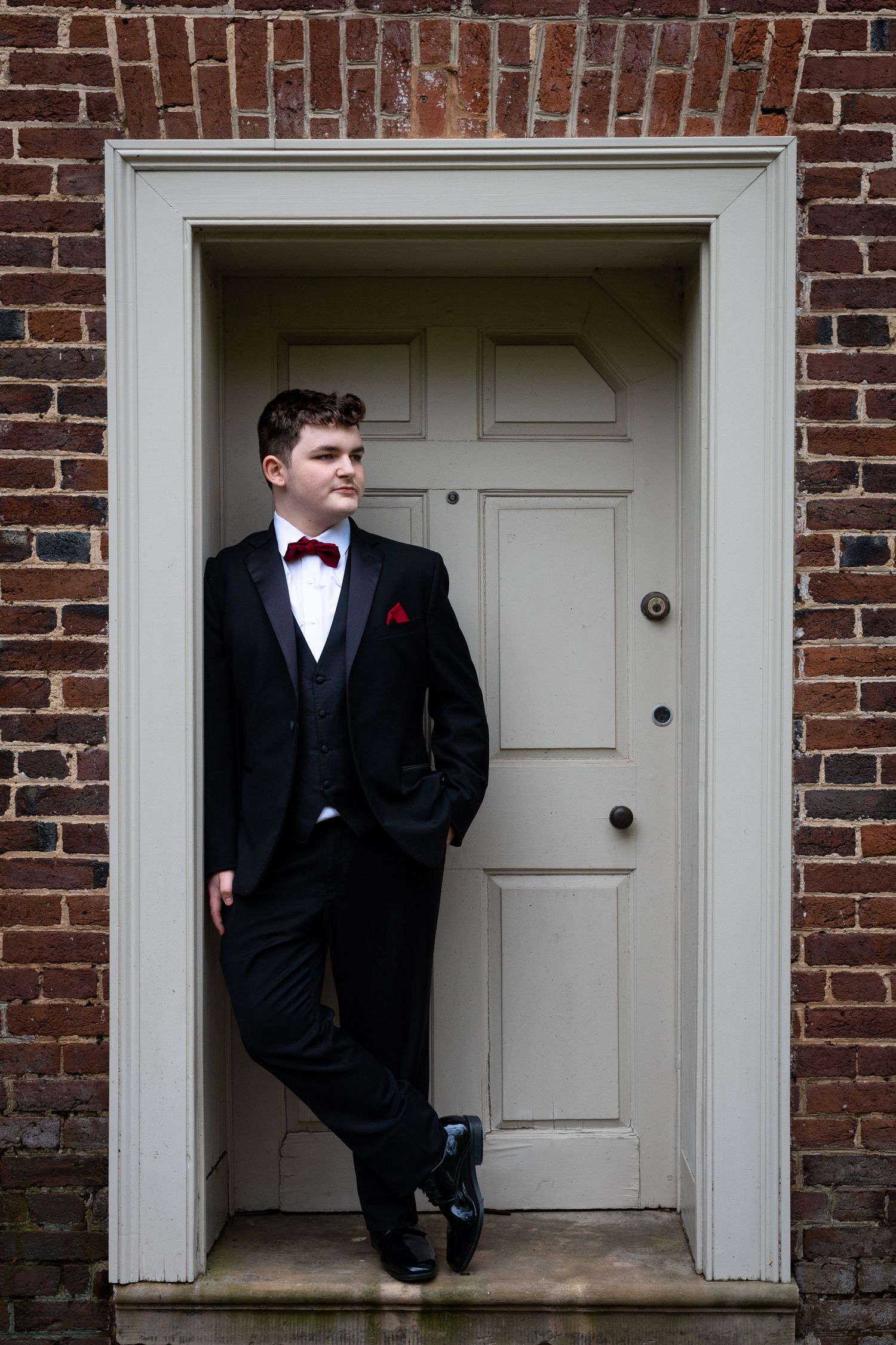 Formal portrait in doorway featuring person in black tuxedo leaning against white frame.