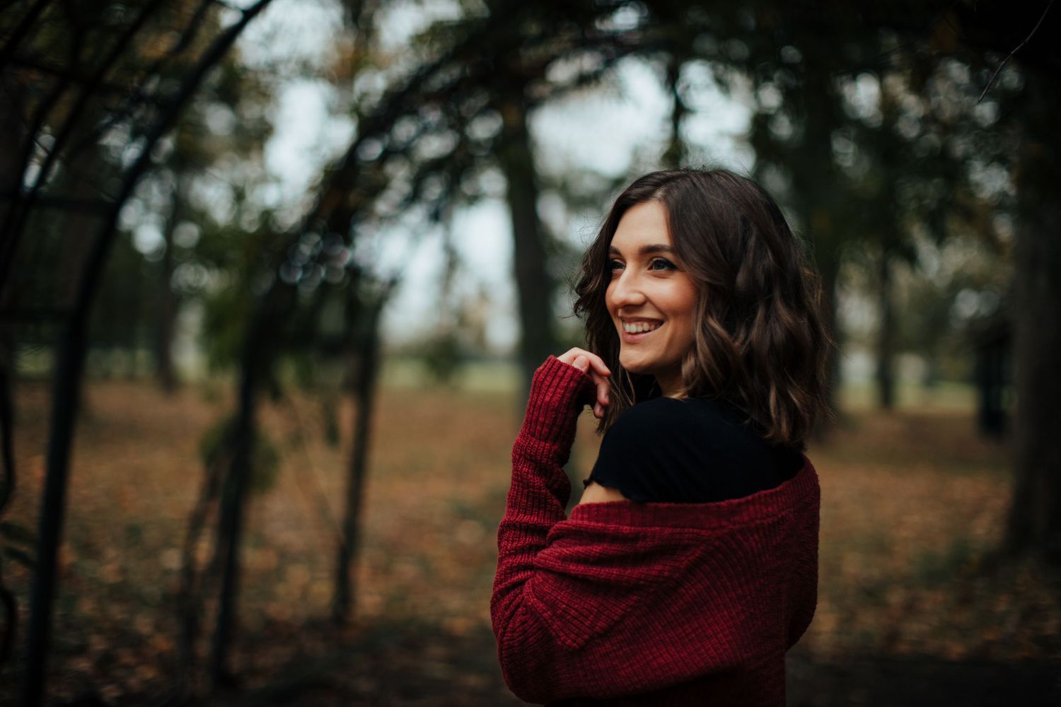A person in a burgundy sweater smiles while standing in an autumn forest.