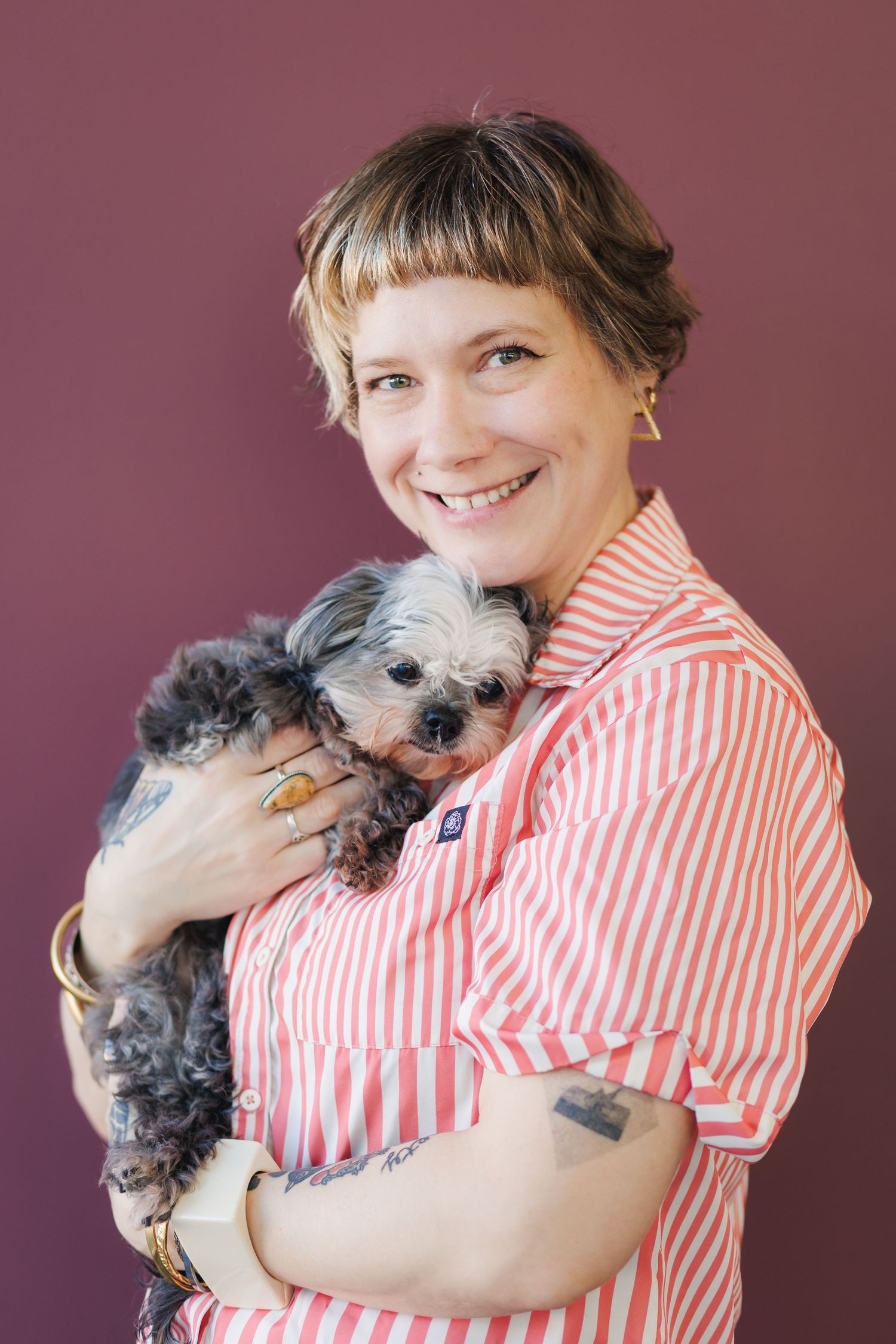 Person wearing red and white striped shirt hugs and cuddles with small gray scruffy dog in studio setting.
