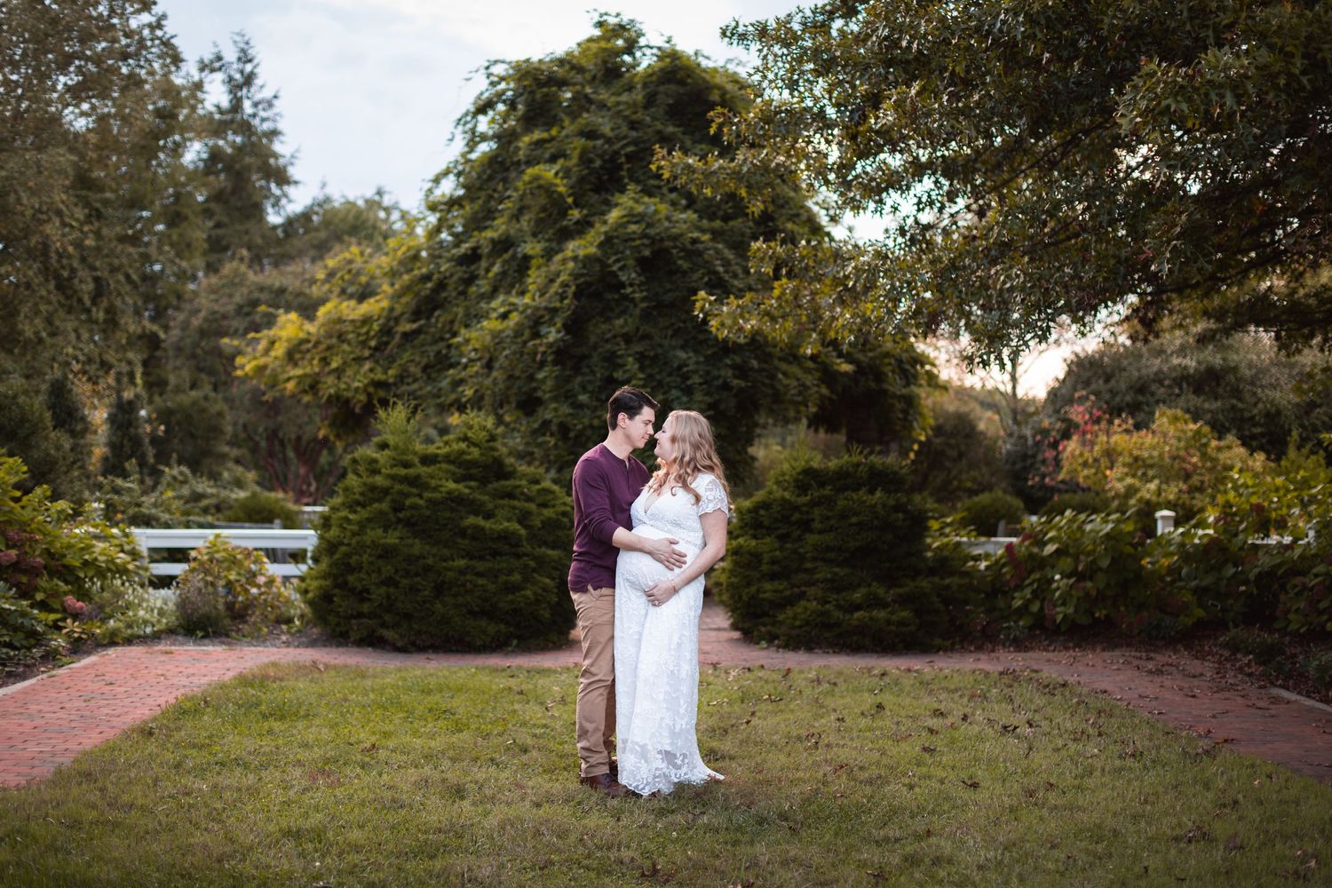 A romantic maternity photo of a couple embracing in a garden with manicured hedges.