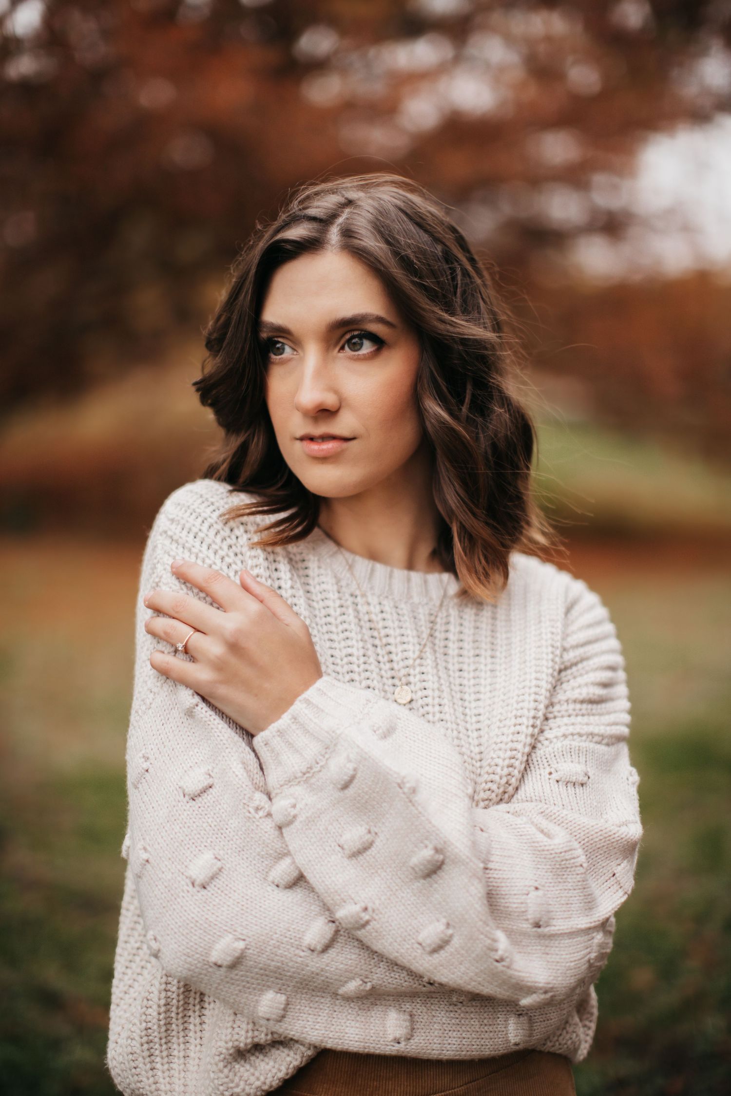 Portrait in cream knit sweater against blurred fall foliage background.