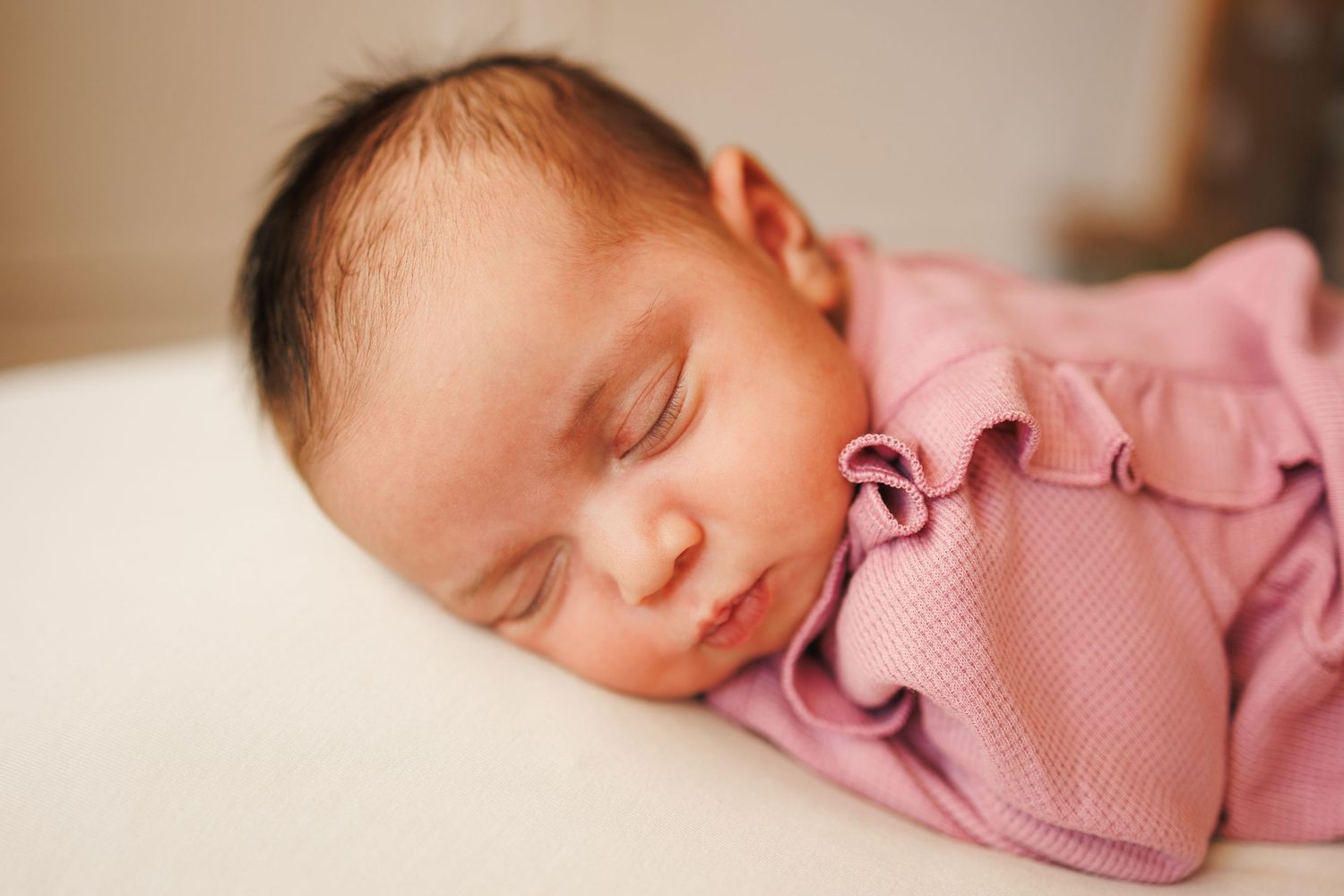 Newborn baby sleeping peacefully in soft pink clothing against a neutral background.