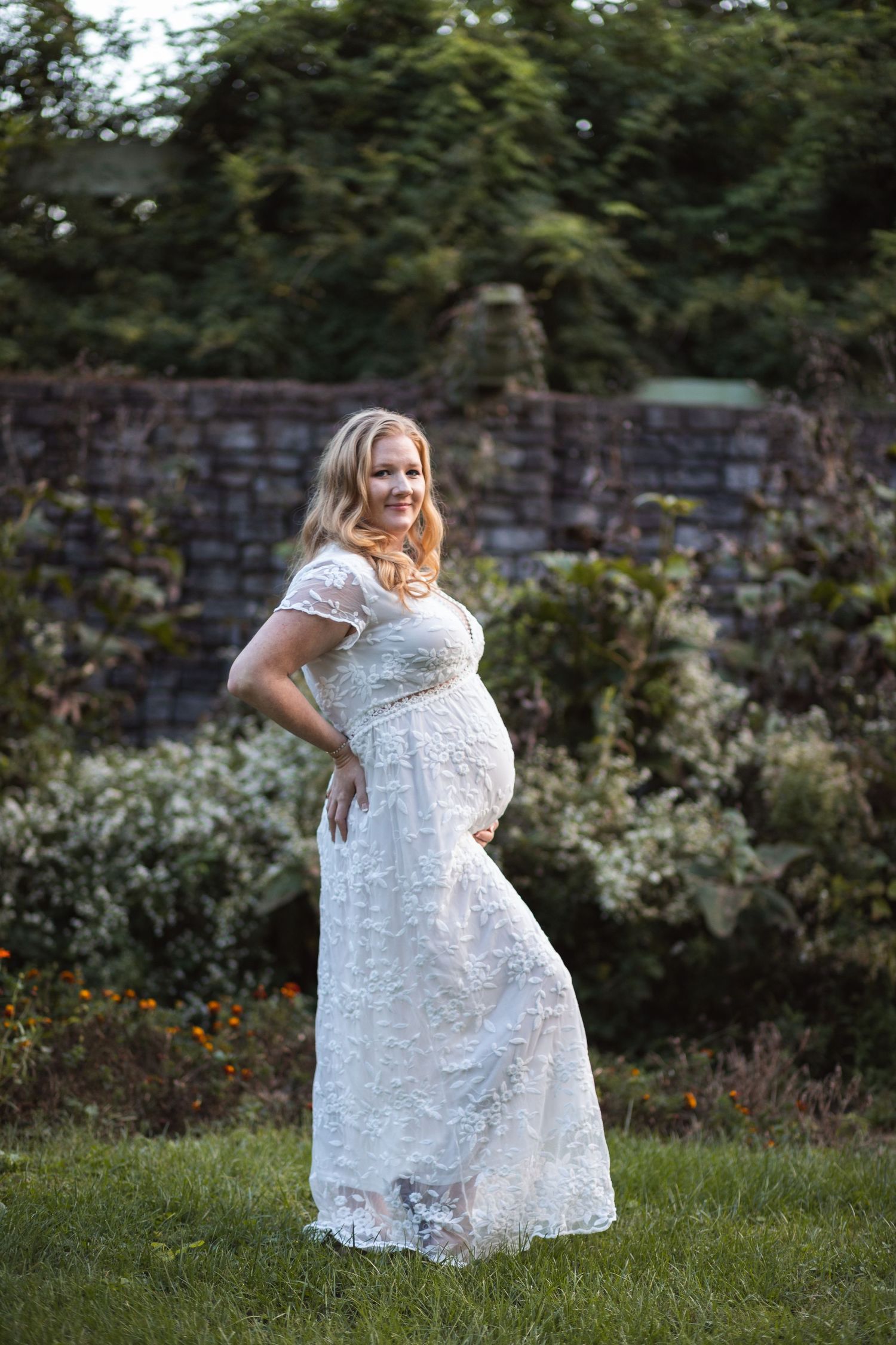 A maternity portrait featuring a woman in a white lace dress posing by a stone wall.