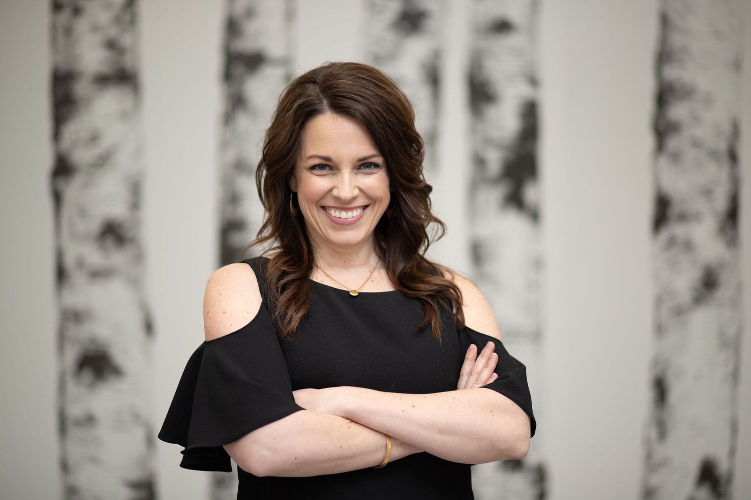 A woman in a black cold-shoulder dress smiles while standing with crossed arms against a light background.