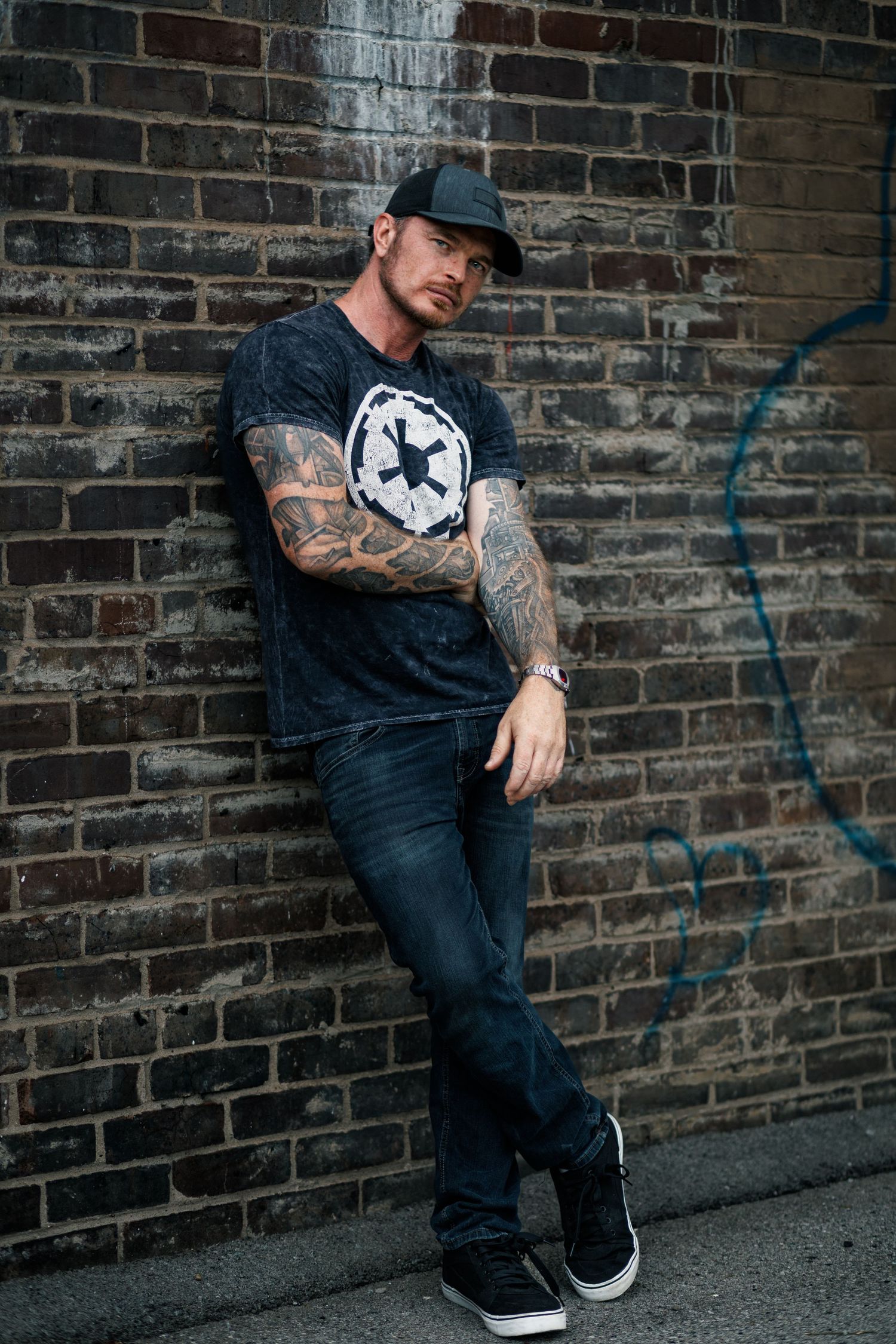Tattooed person in casual streetwear poses against a dark brick wall in three similar urban portraits.