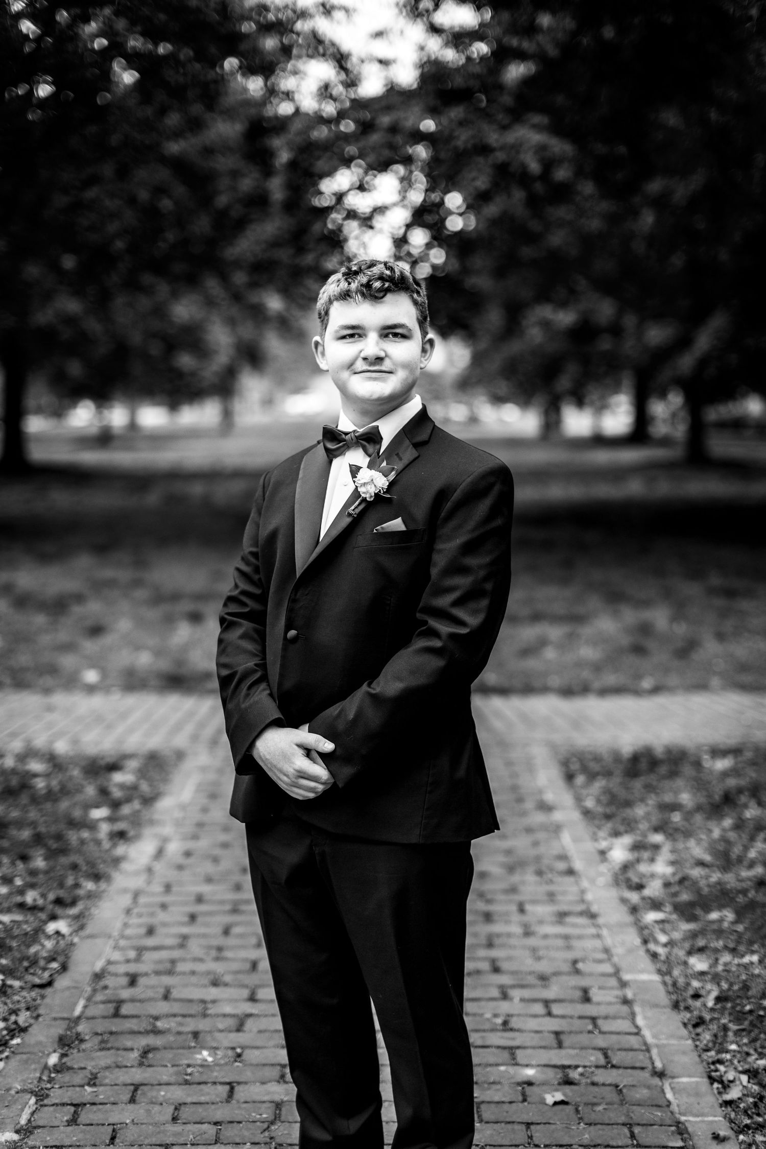 Black and white portrait of someone in a formal tuxedo standing on a brick path with trees in the background.